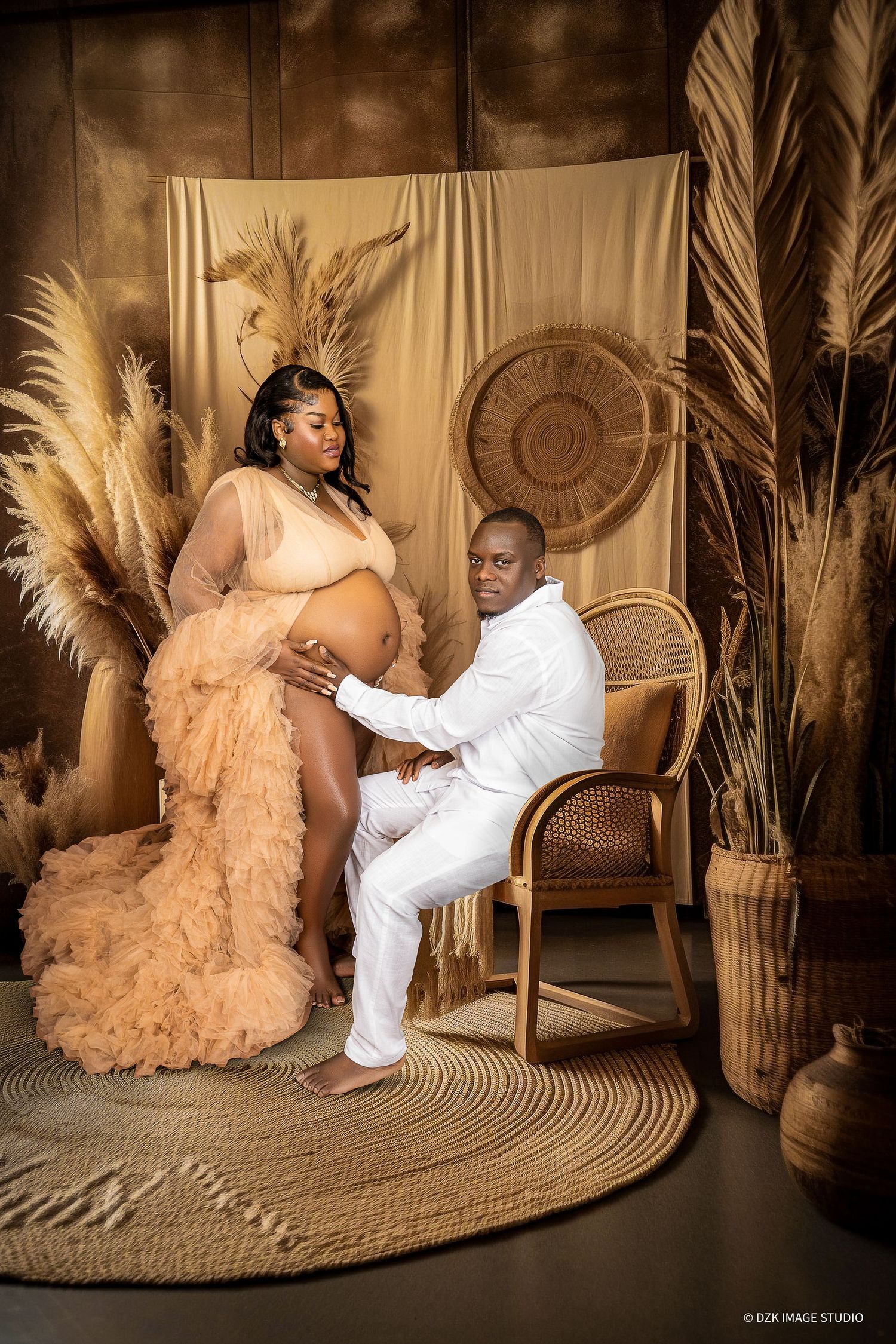 Expectant couple in peach and white outfits pose in a boho studio with pampas grass, a rattan chair and woven decor.