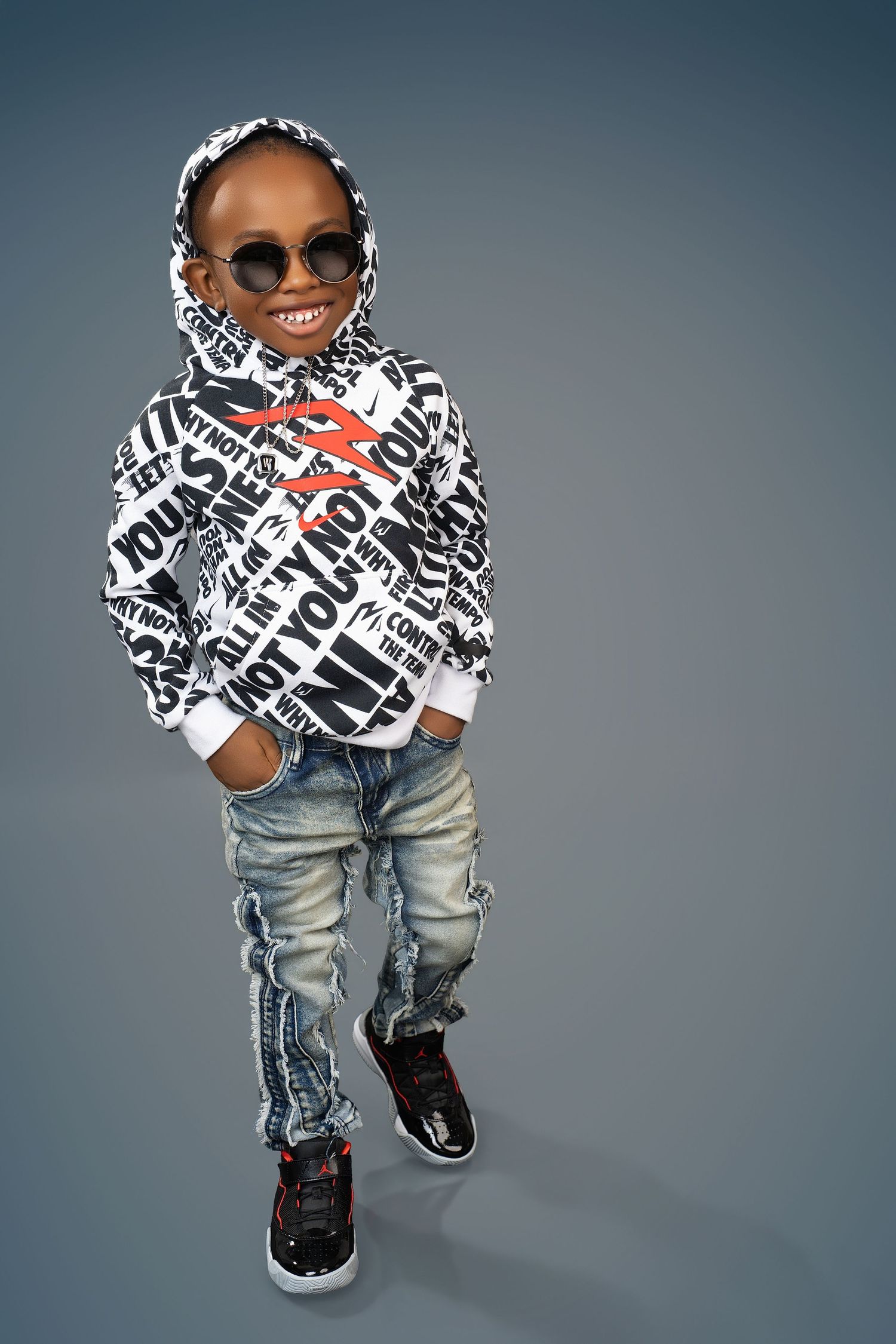 Smiling boy in round sunglasses, a black and white graphic hoodie, distressed jeans and black sneakers posing against a gray backdrop.