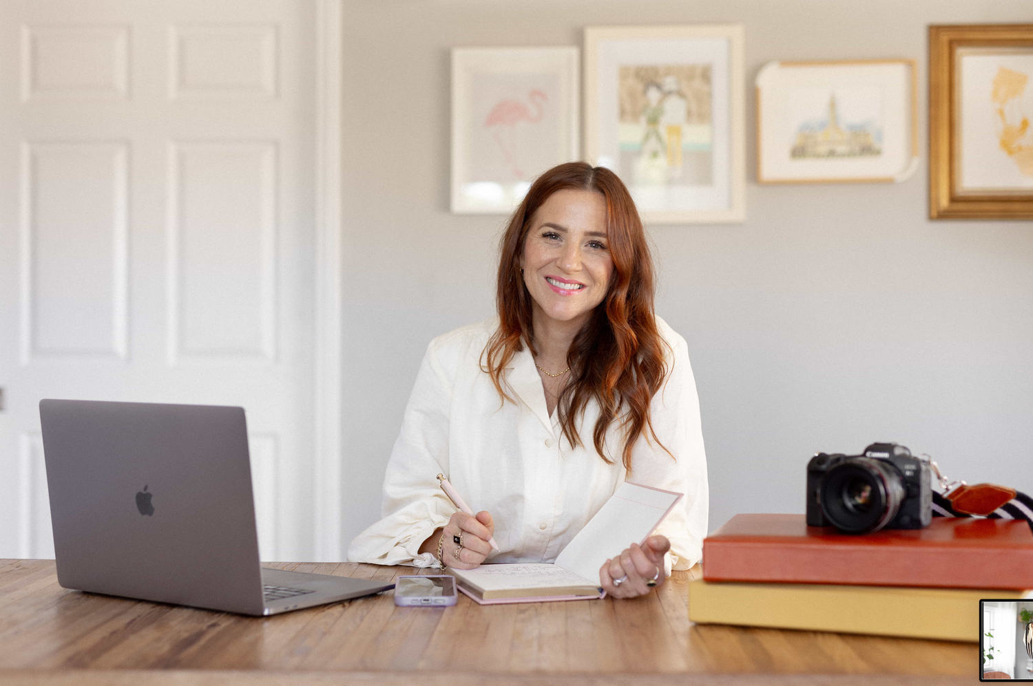 Smiling woman with red hair sits at wooden desk with laptop, notebook, and camera in bright home office.