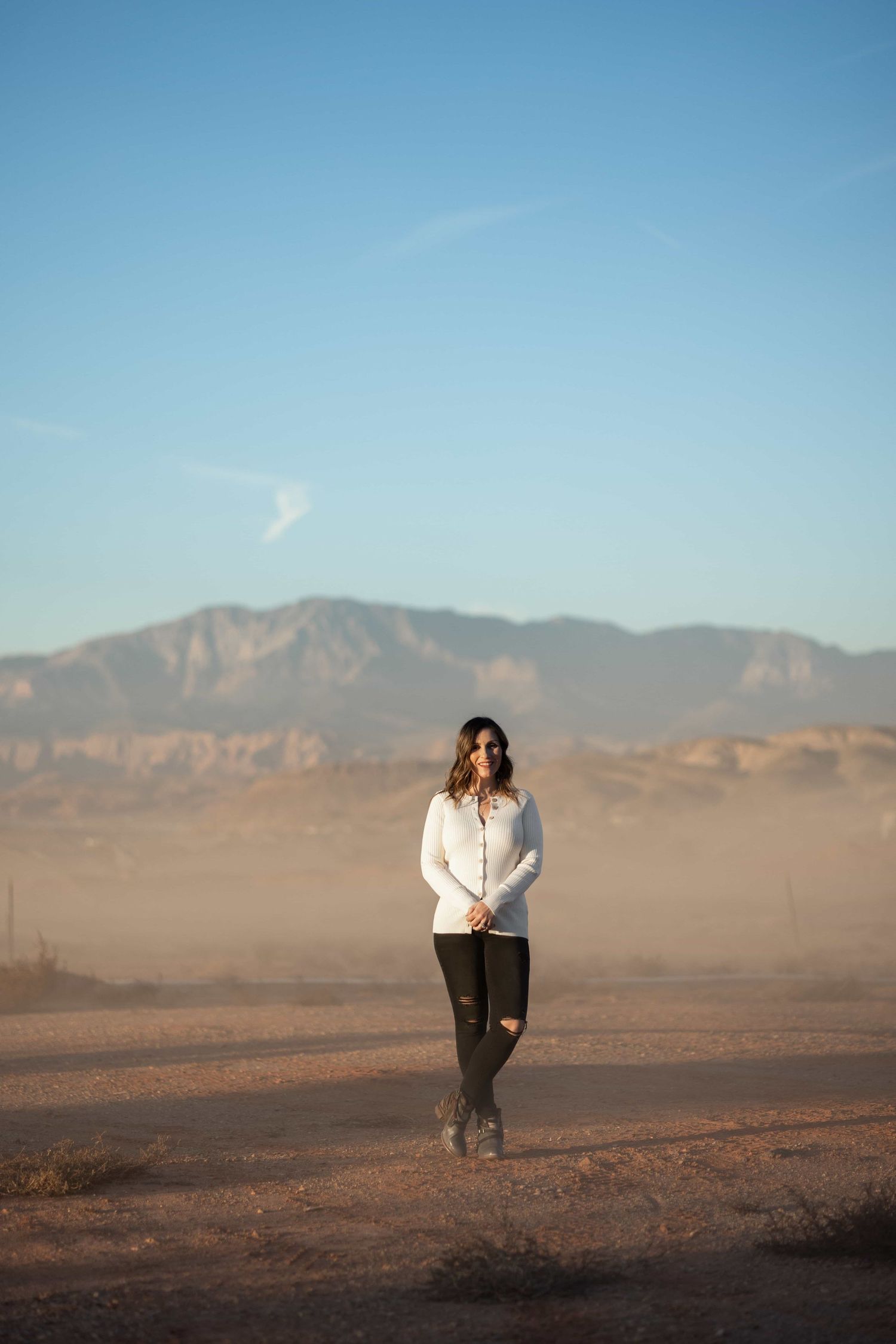 Woman in white top and black jeans stands in dusty desert landscape with mountains and blue sky behind her.
