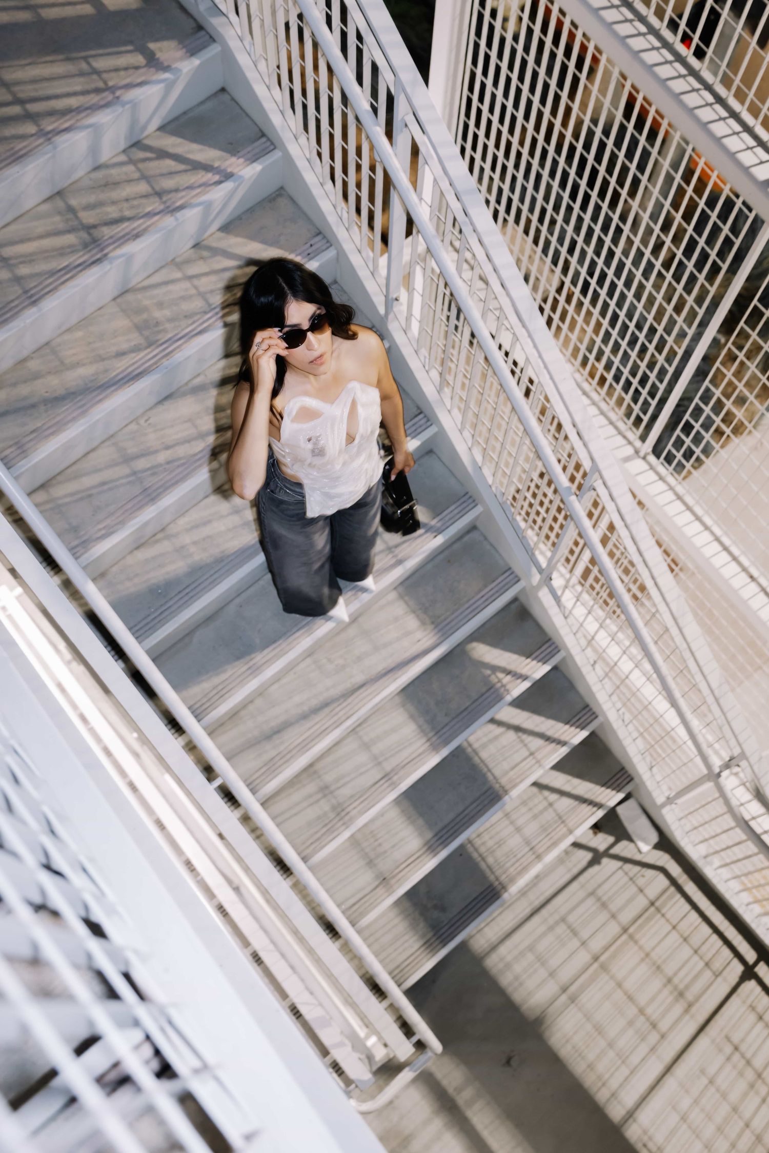 Woman in sunglasses and casual outfit descending metal outdoor stairs, viewed from above.