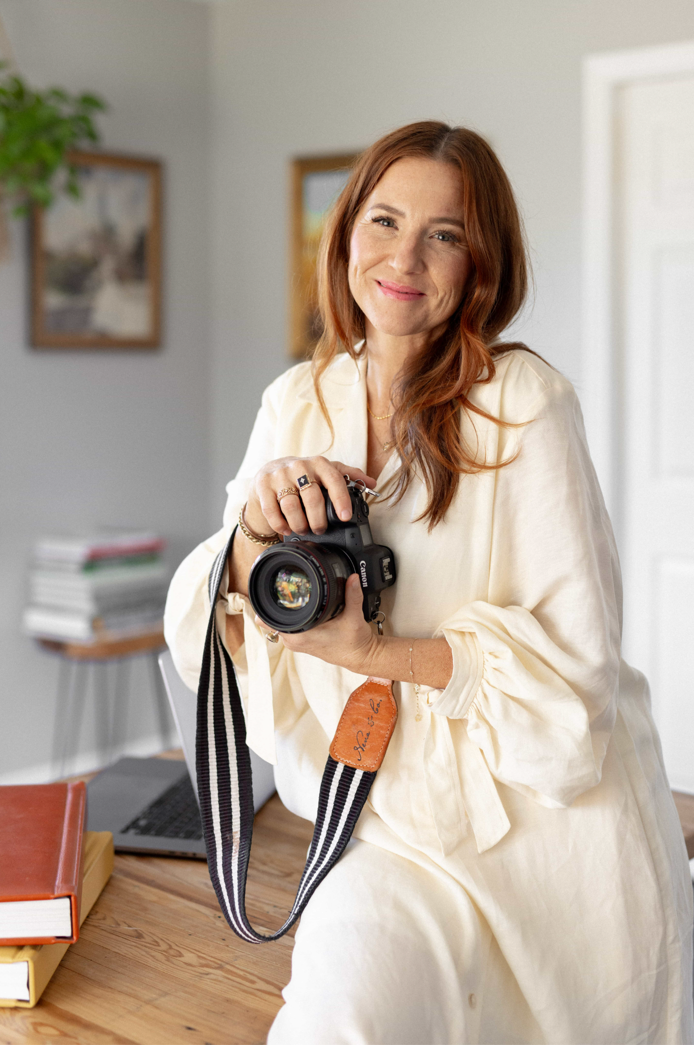 Woman in cream outfit holding a DSLR camera with striped strap, smiling in a bright home studio setting.