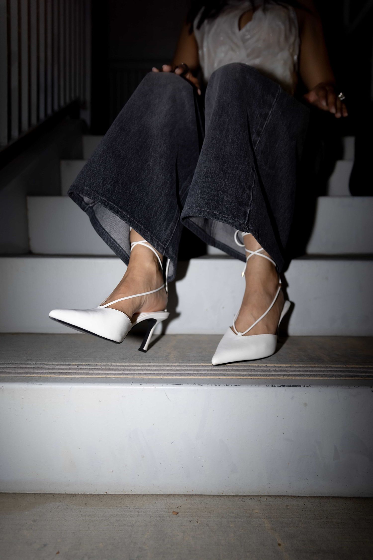 Person sitting on white steps wearing dark flare jeans and white pointed-toe slingback heels with thin ankle straps.