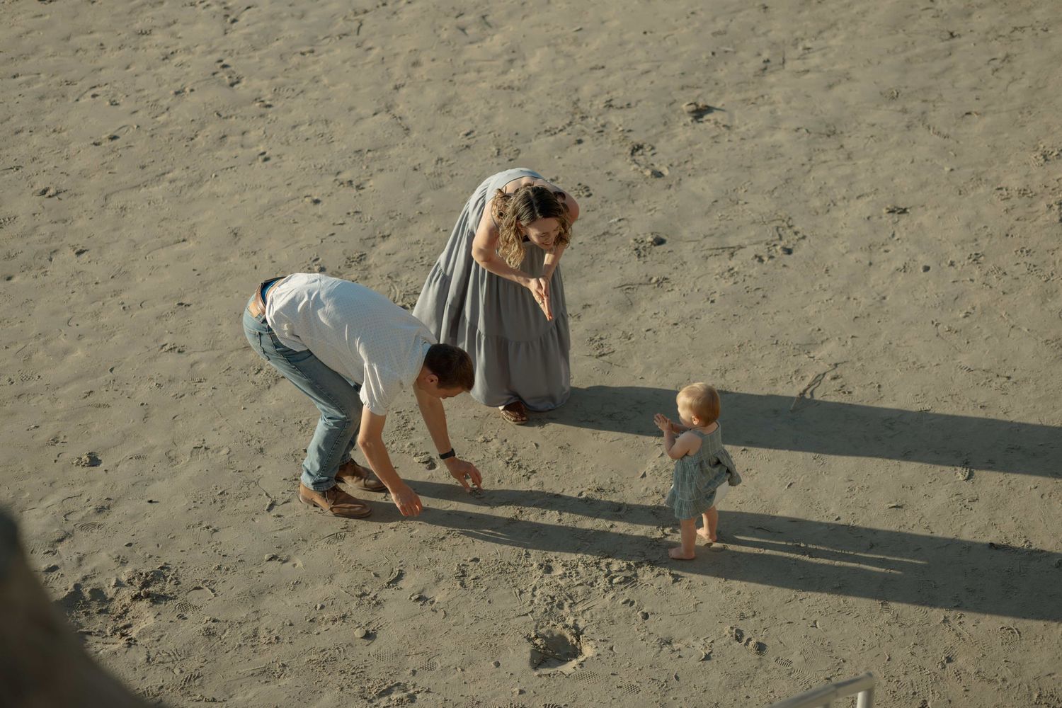Two adults bending down to interact with a toddler on a sandy beach, captured from above.