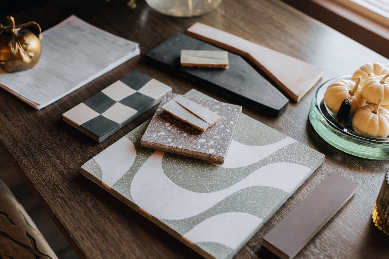 Decorative tile samples in checkered,terrazzo,and wavy patterns displayed on a rustic wooden table with golden accents.
