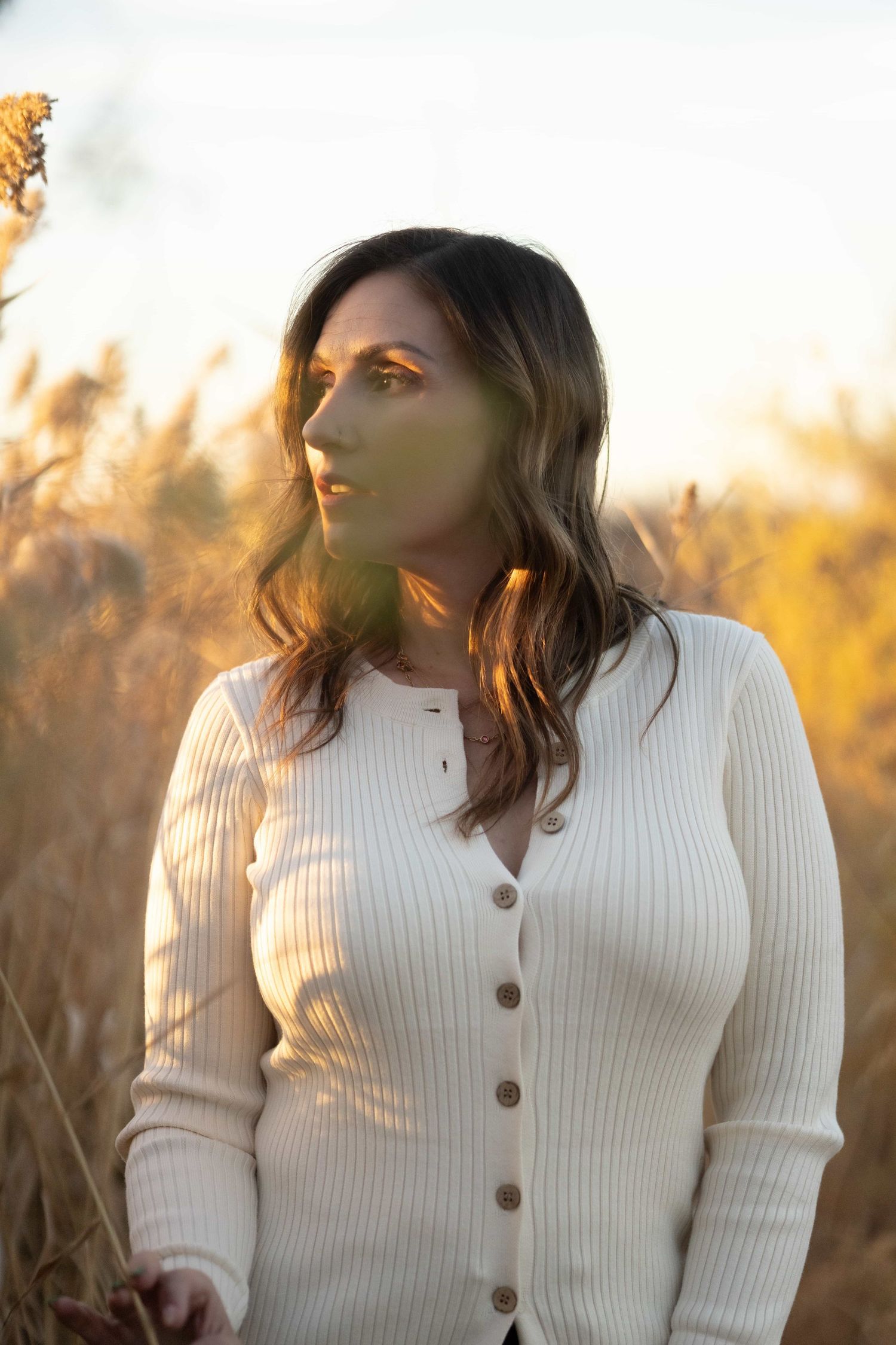 Woman in white ribbed cardigan standing in golden field during sunset with warm backlight.