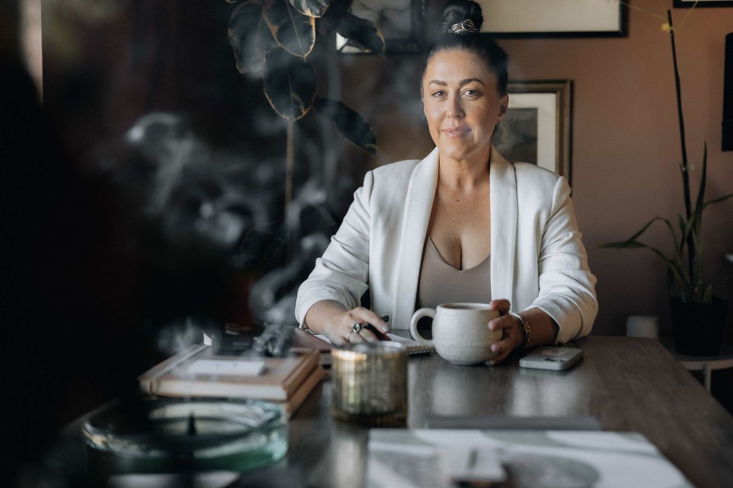 Woman in white blazer holding coffee mug at table with smoke rising, moody cafe atmosphere.