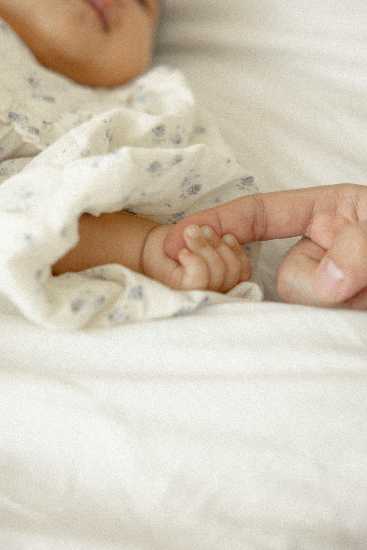 Newborn baby grasping adult finger while sleeping on white bedding in floral outfit.