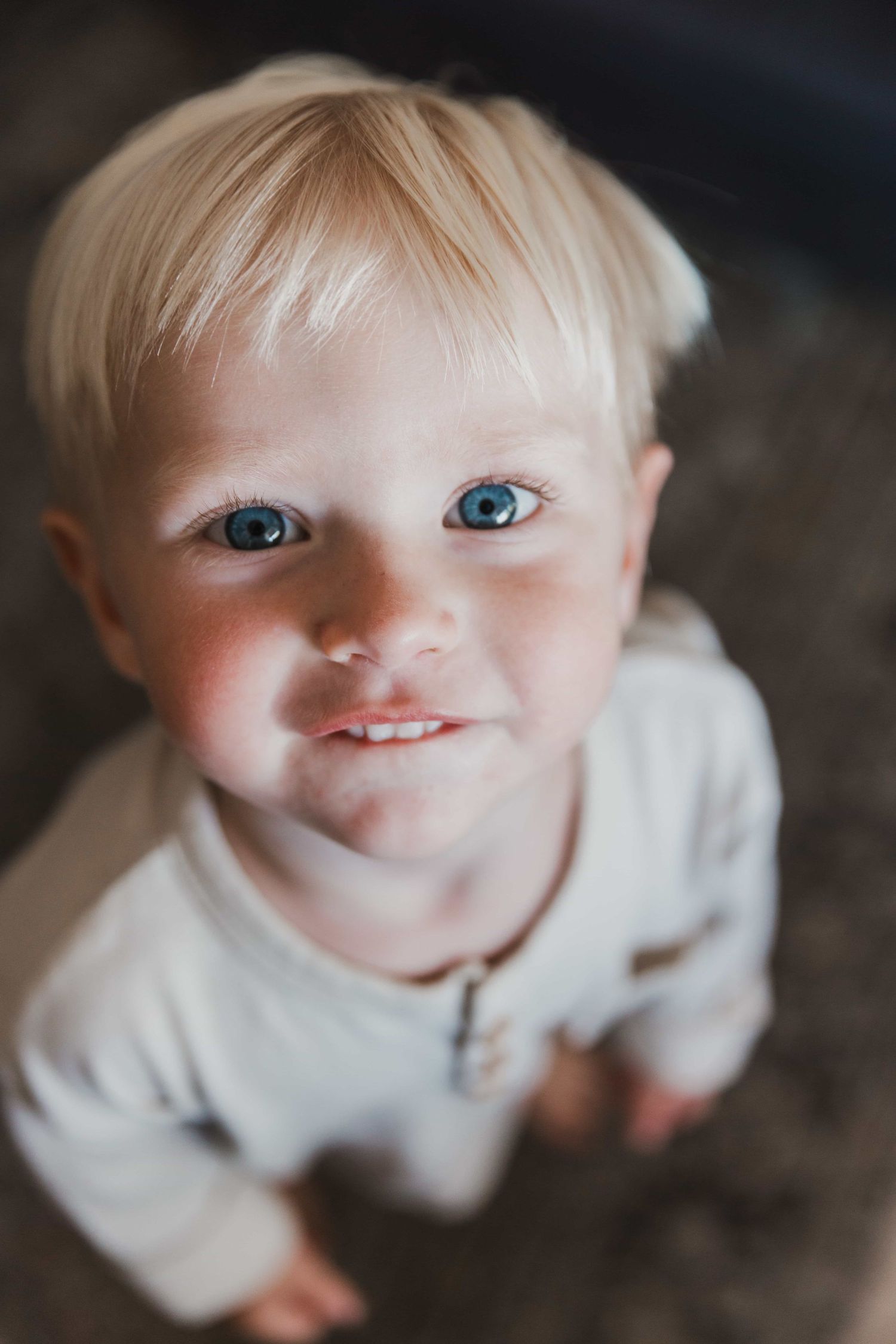 Adorable blonde toddler with blue eyes smiling up at camera in soft indoor lighting.