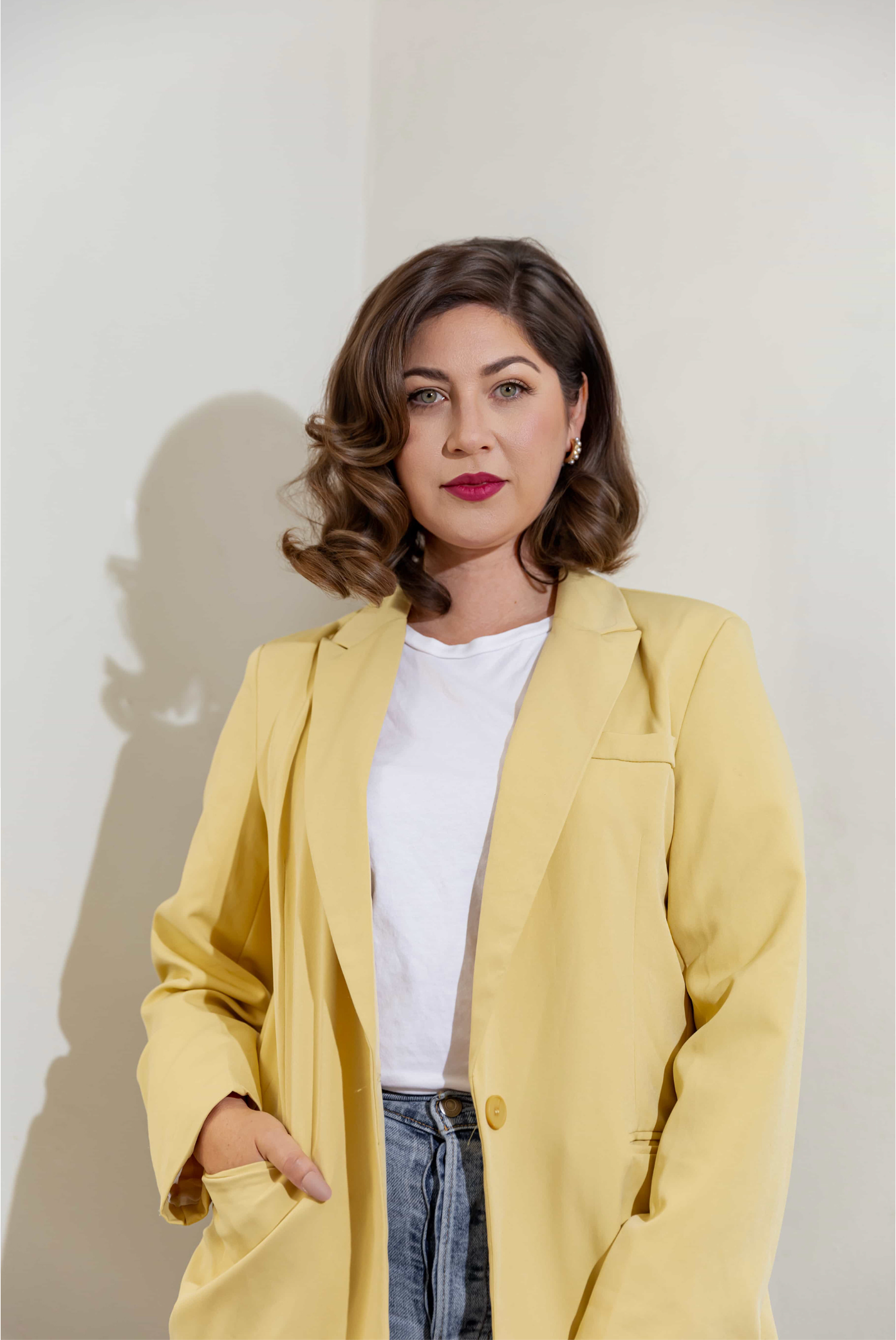Woman in yellow blazer and white top with jeans, posing against light wall with red lips and wavy hair.