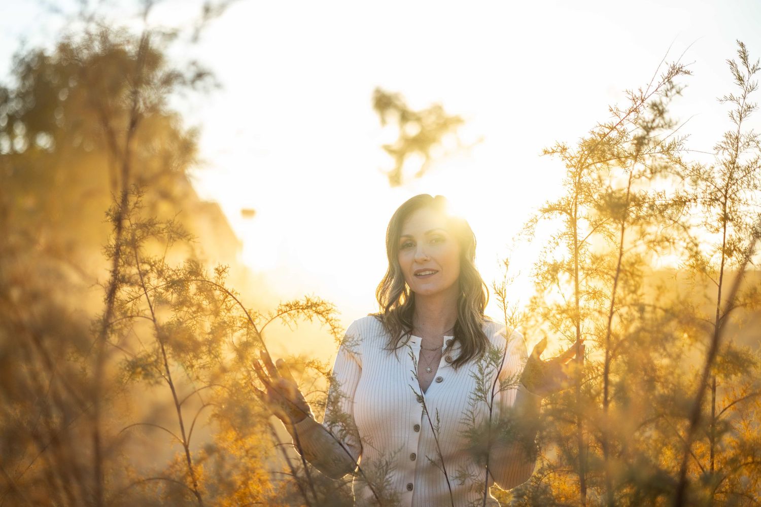 Woman smiling in golden sunset light surrounded by tall wild grasses and plants in a warm outdoor field.