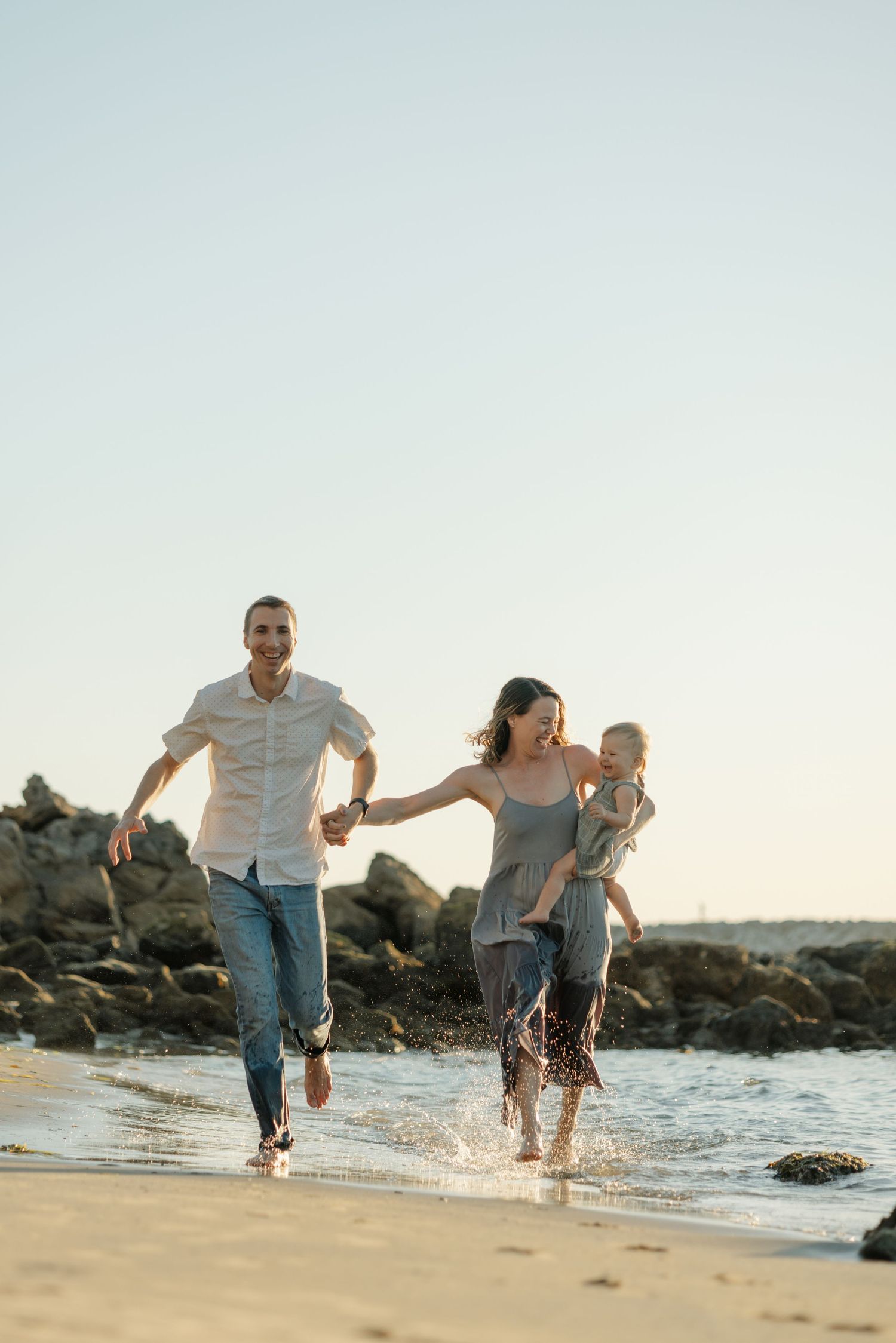 Happy family running along the beach shoreline, parents holding hands with young child, splashing in shallow waves at sunset.