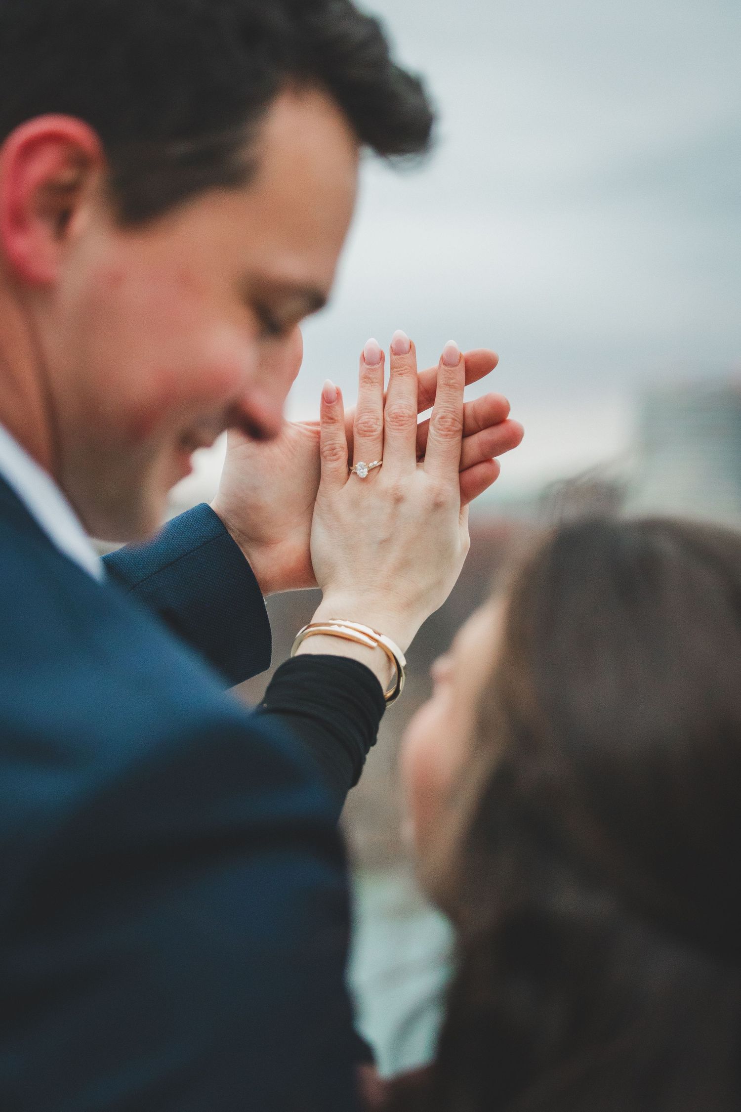 Two hands clasped together showing off engagement ring against a blurred background, wearing a dark blue suit sleeve.