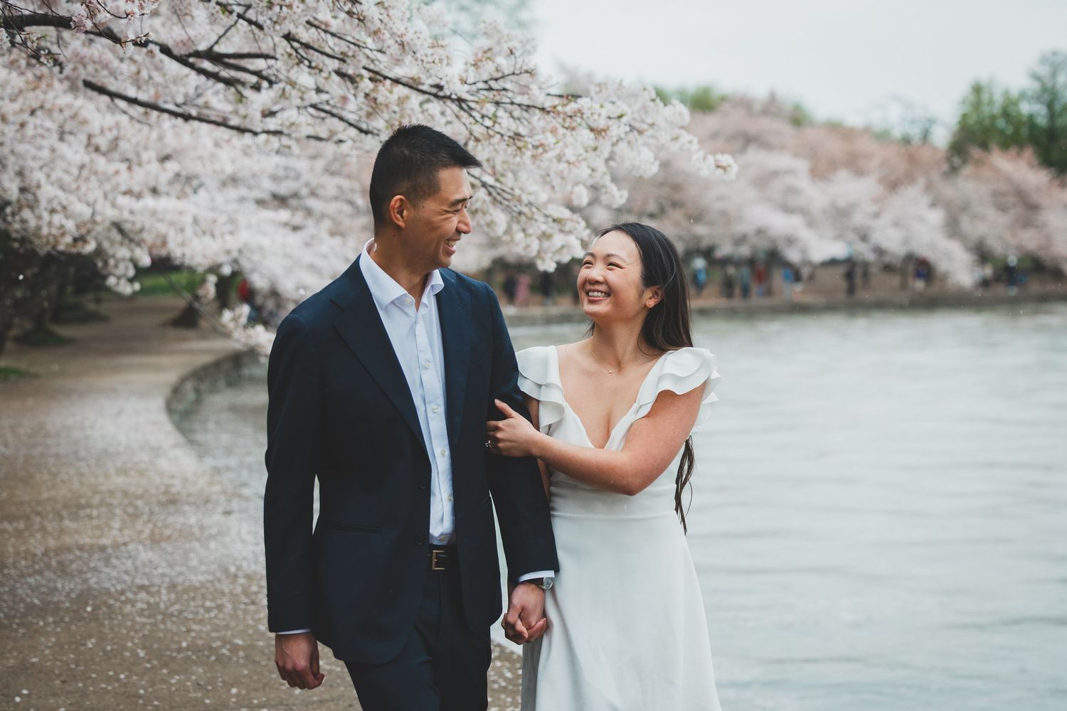 Couple smiling and walking together under blooming cherry blossom trees near a waterfront in spring.