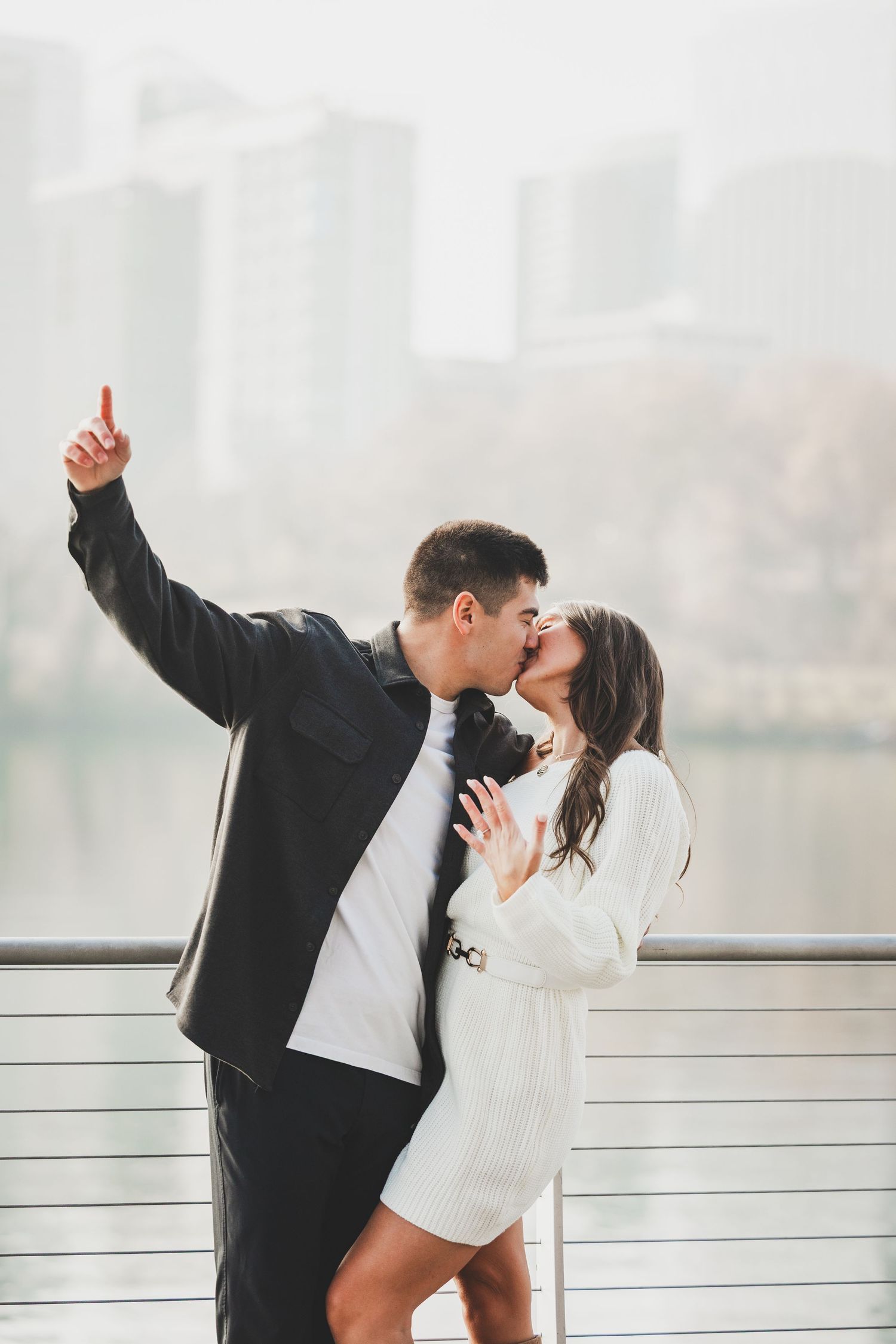 A romantic couple shares a tender kiss while standing against a railing with a city skyline in the background.