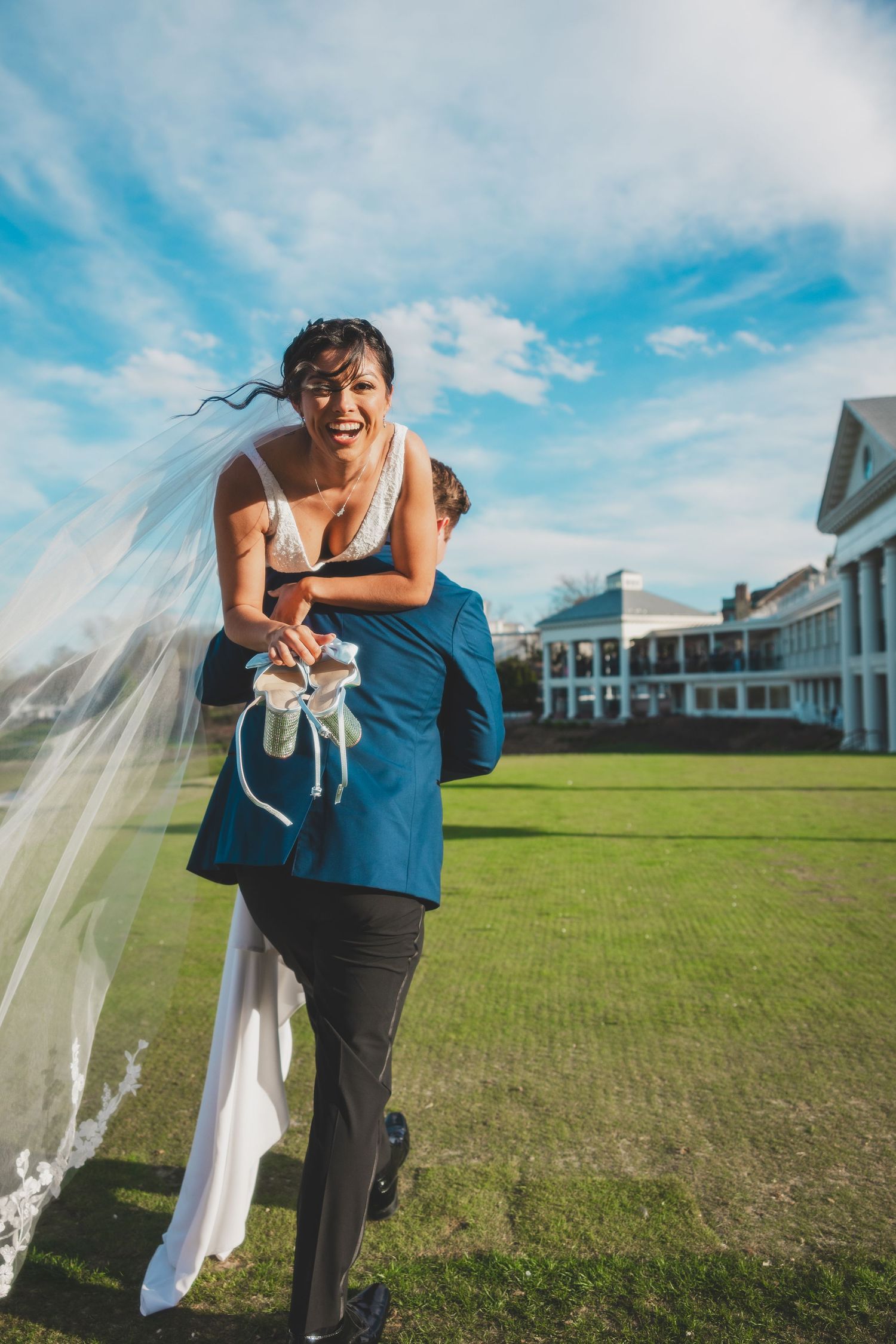 Playful piggyback wedding photo on a sunny day with a mansion in the background.