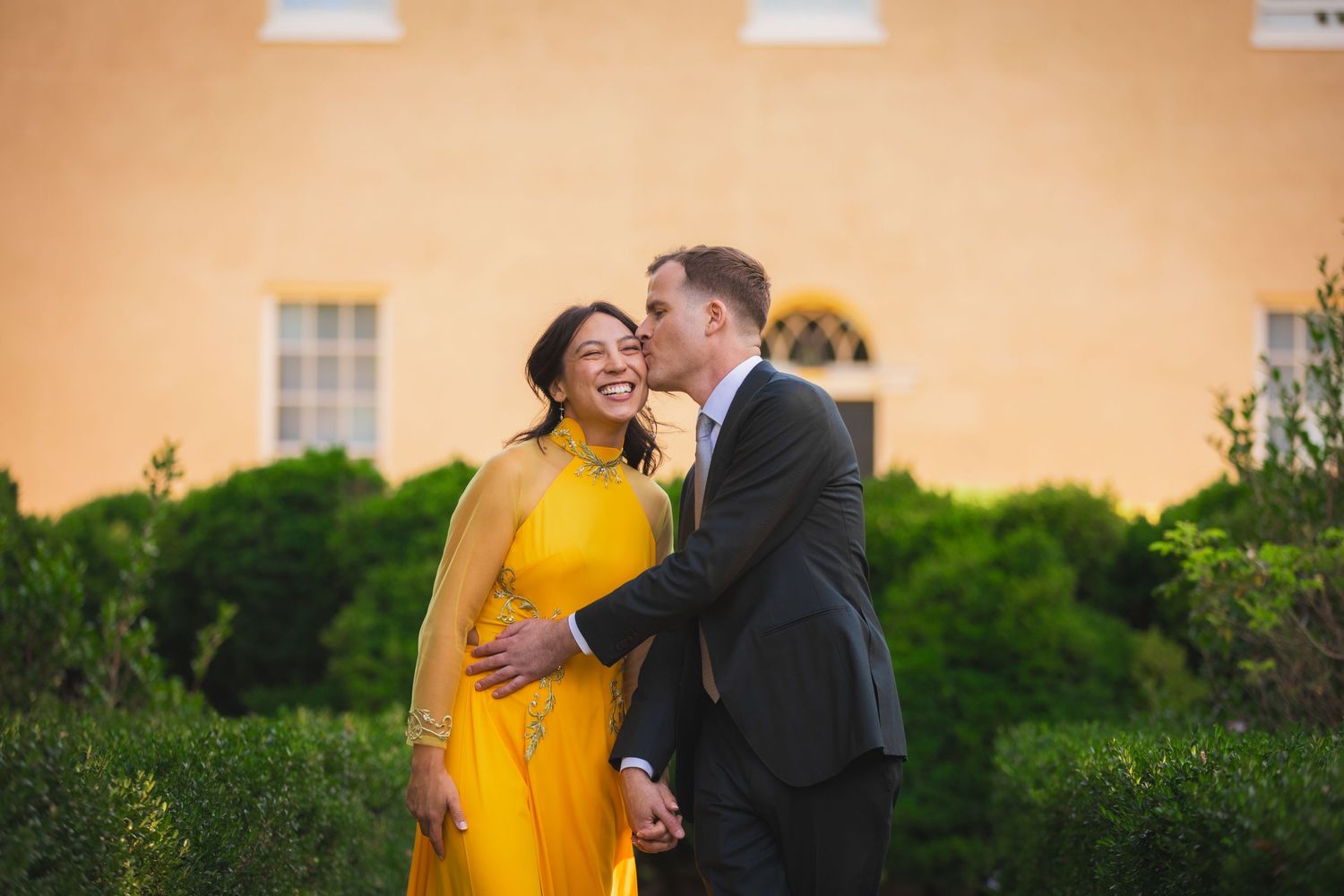 Romantic outdoor portrait of couple laughing together against orange building and garden backdrop.