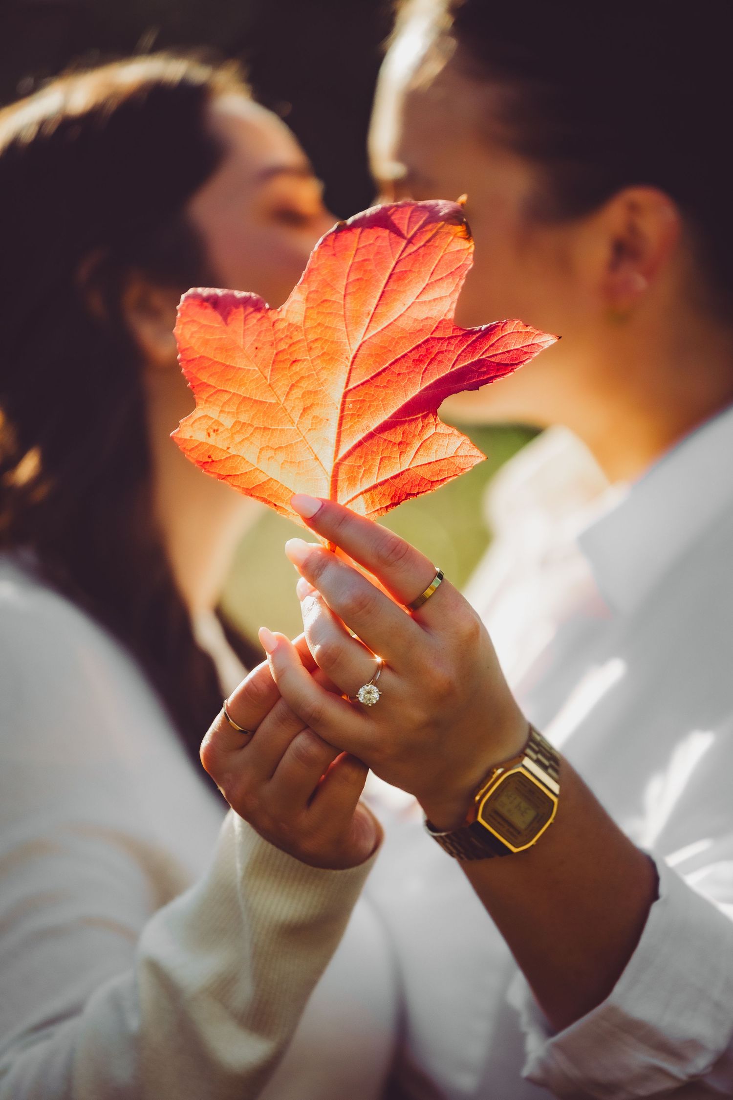 An engaged couple holding a vibrant orange autumn maple leaf against a blurred background in soft natural light.