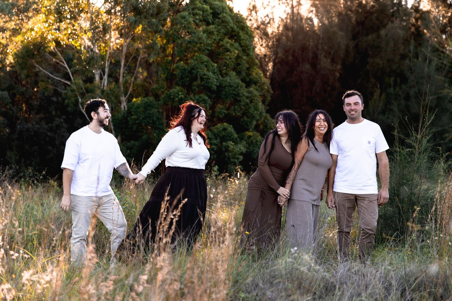 Five adults holding hands and laughing as they walk through long grass in front of tall trees