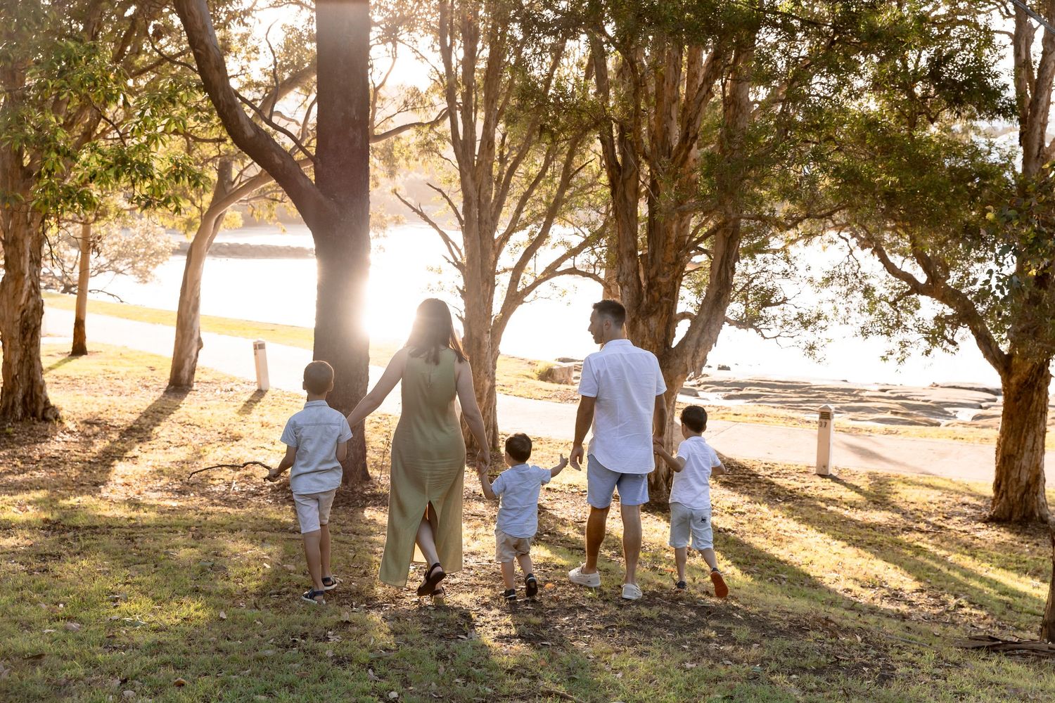 Family of five walking hand in hand under trees towards the water at sunset