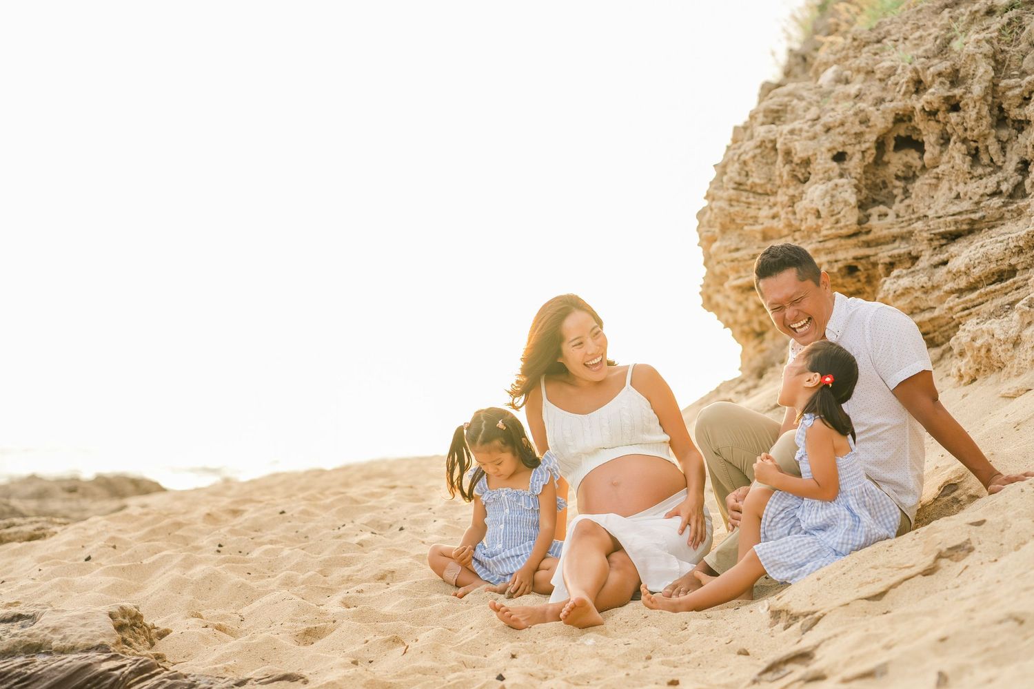 A family sits together on a sandy beach at sunset with cliffs in the background. Oahu Family Photographer
