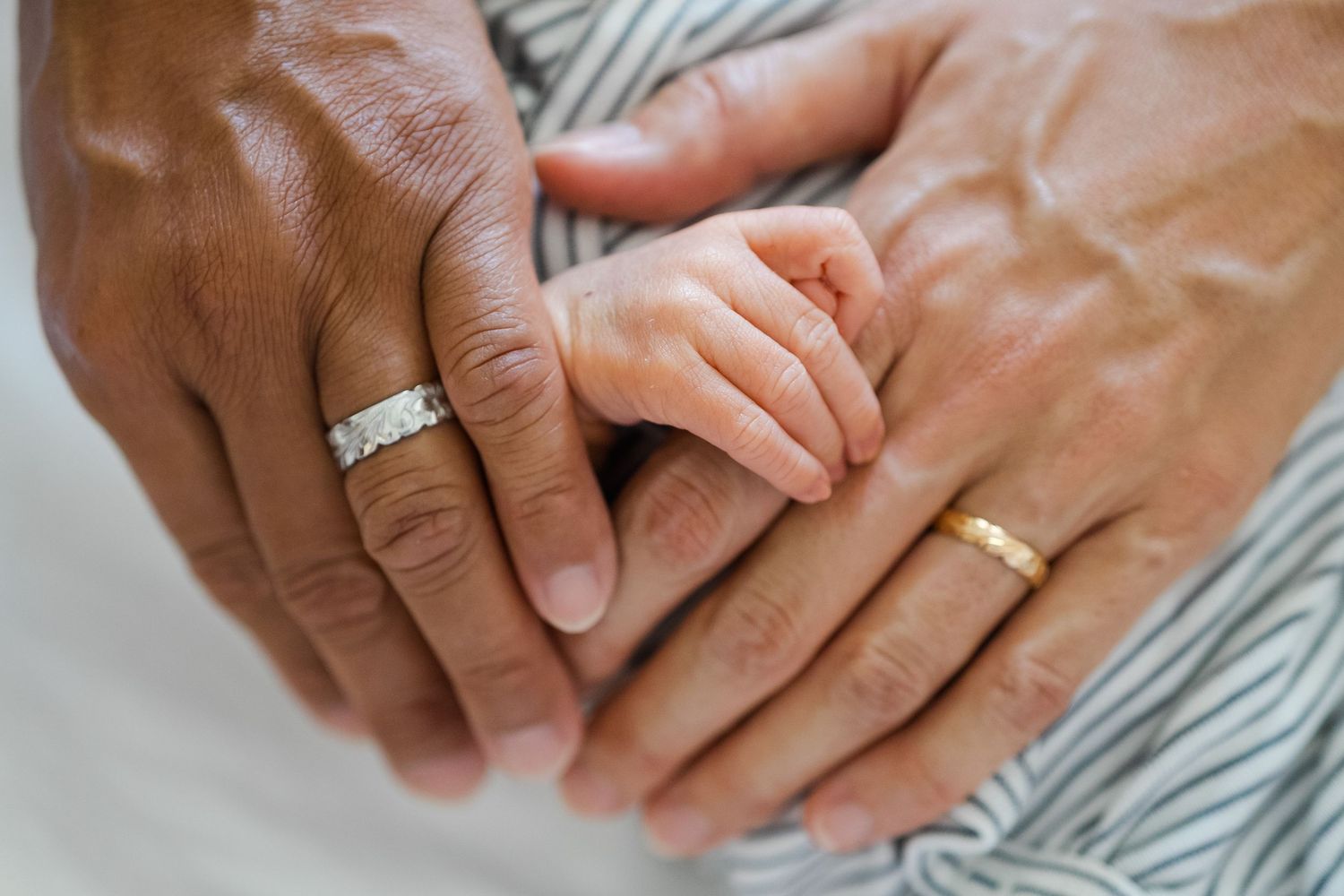 Close-up view of two hands wearing wedding bands against a light fabric background. Oahu Newborn Photographer