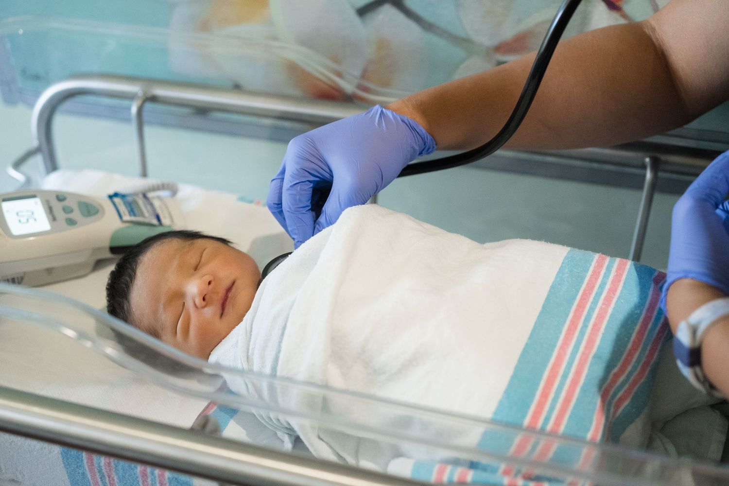 Medical staff with gloved hands examines a sleeping newborn infant wrapped in a blanket in hospital bassinet. Oahu Fresh 48