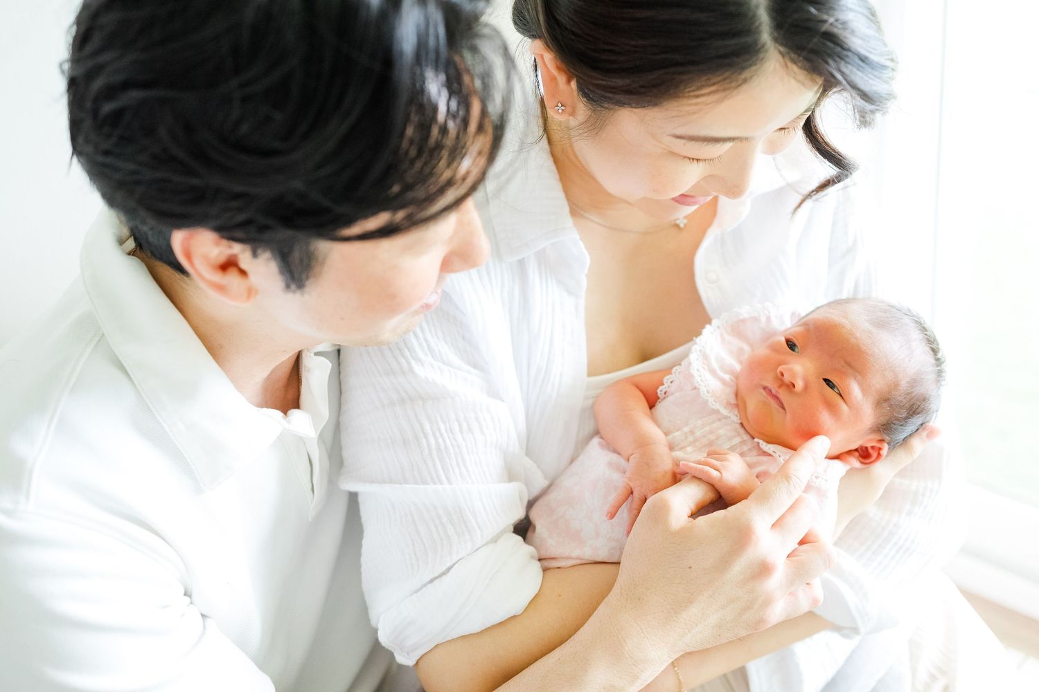 Newborn baby wearing soft pink clothing being cradled gently by two adults in white outfits. Oahu Newborn Photographer