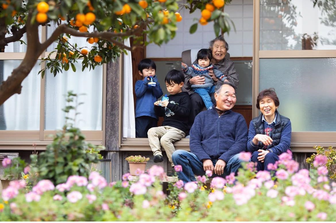 A cheerful group sits on steps beneath a citrus tree, surrounded by blooming pink flowers in a garden setting.