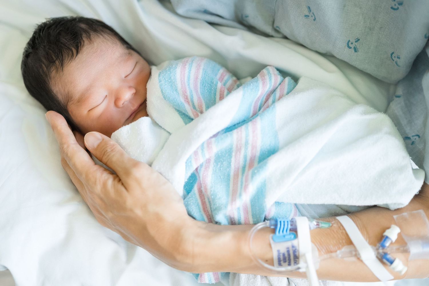 A newborn baby sleeps peacefully while swaddled in a blue and white striped blanket with medical IV attached.