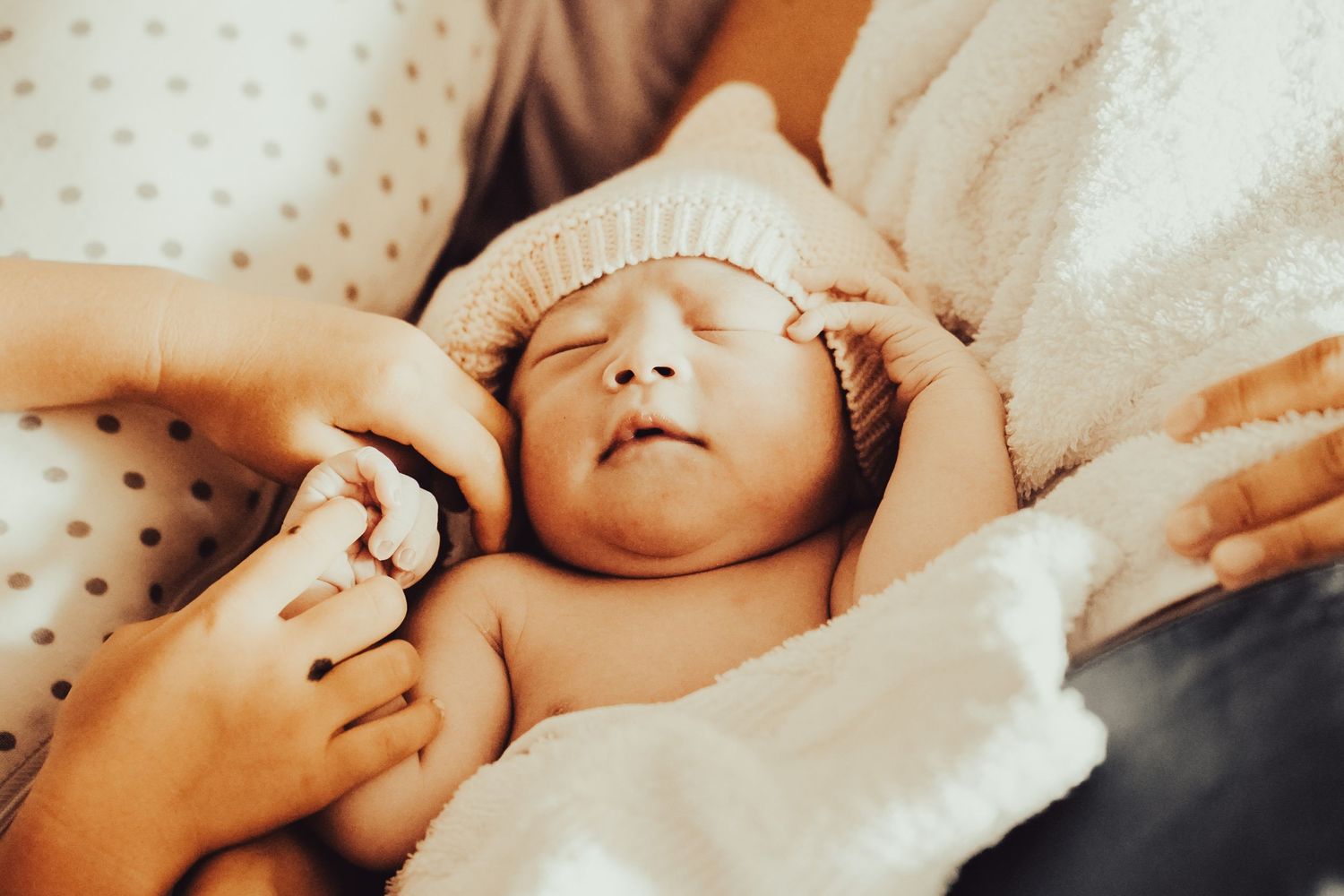 A newborn baby sleeps peacefully while being gently touched by adult hands in a soft, warm setting.