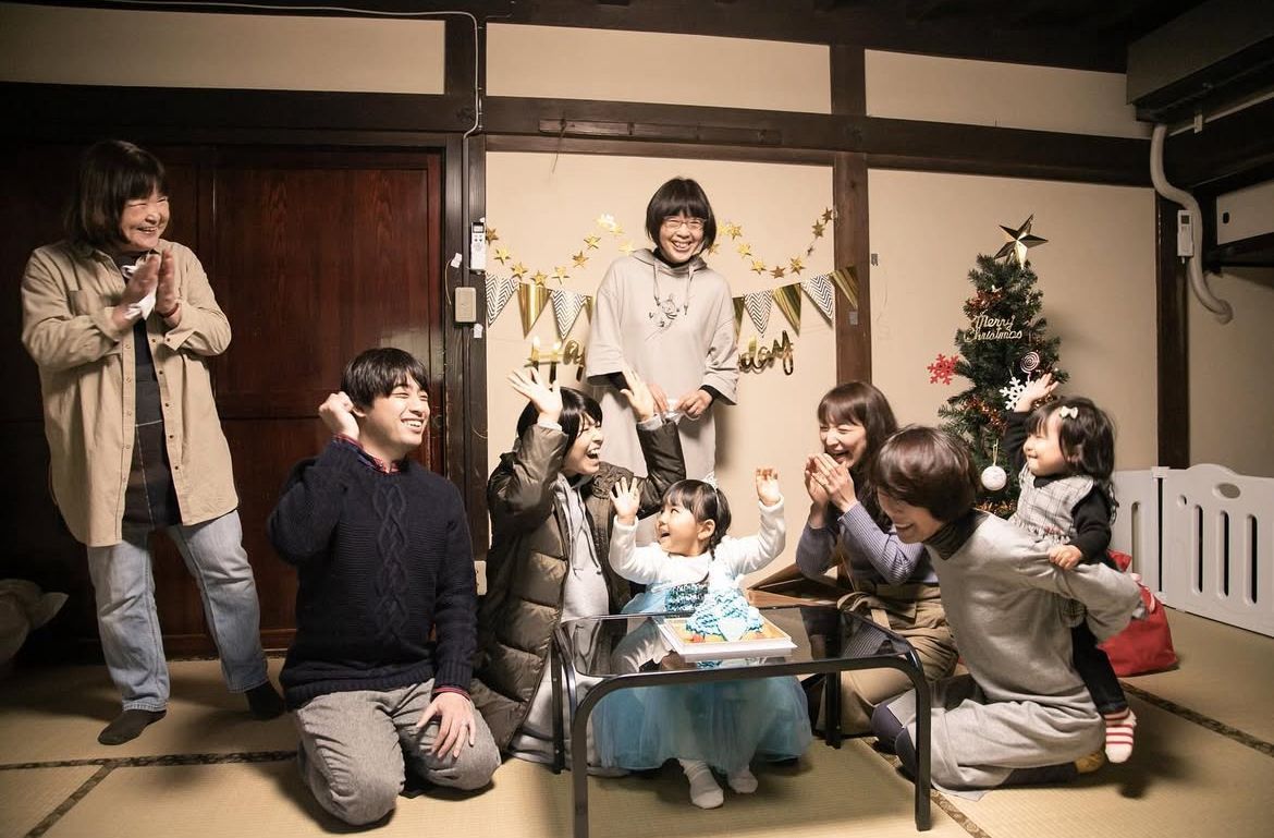 Group gathered in traditional Japanese tatami room celebrating around Christmas tree in casual winter attire.
