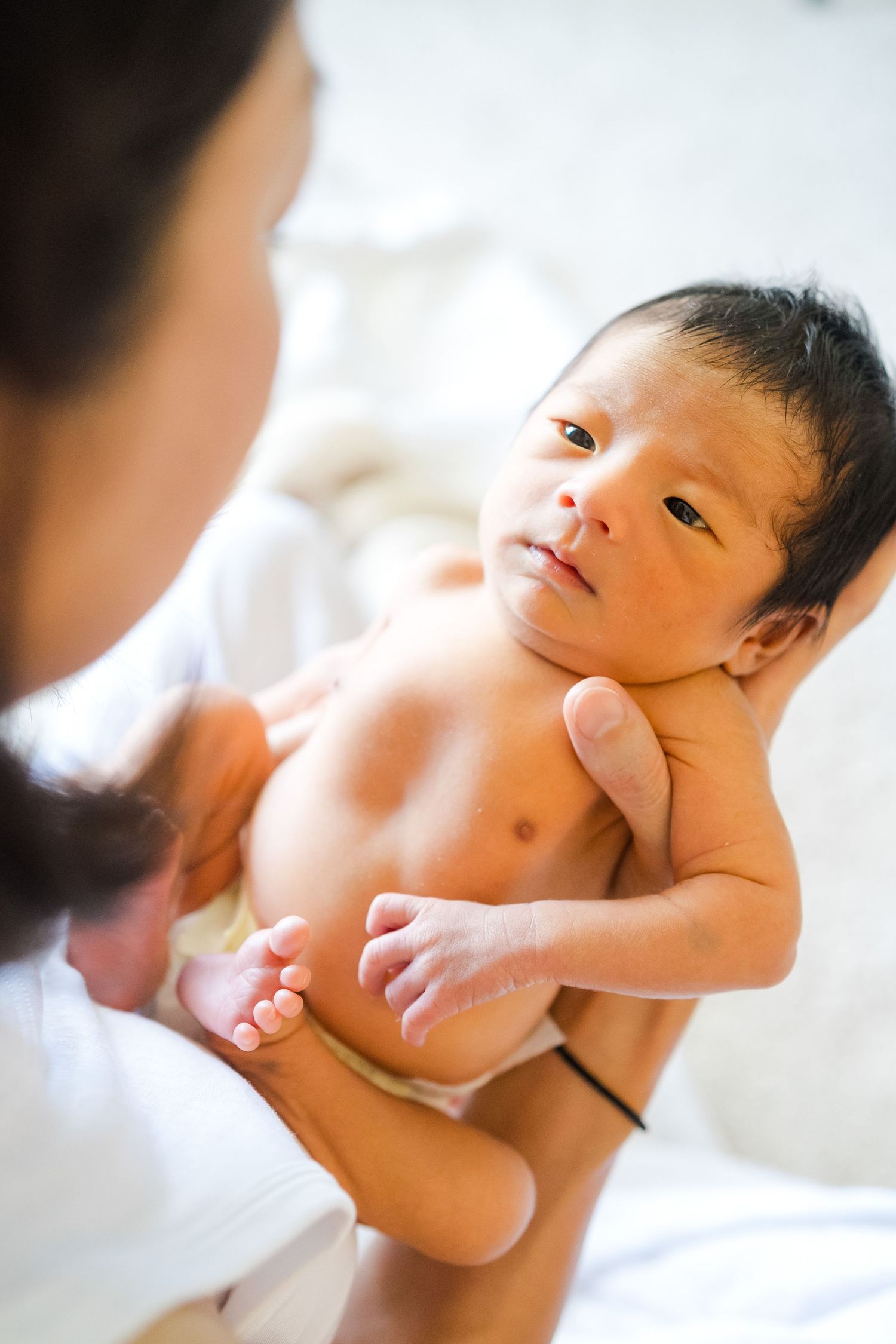 Newborn infant lying in caregiver's hands during a medical examination on white bedding. Oahu Newborn Photographer