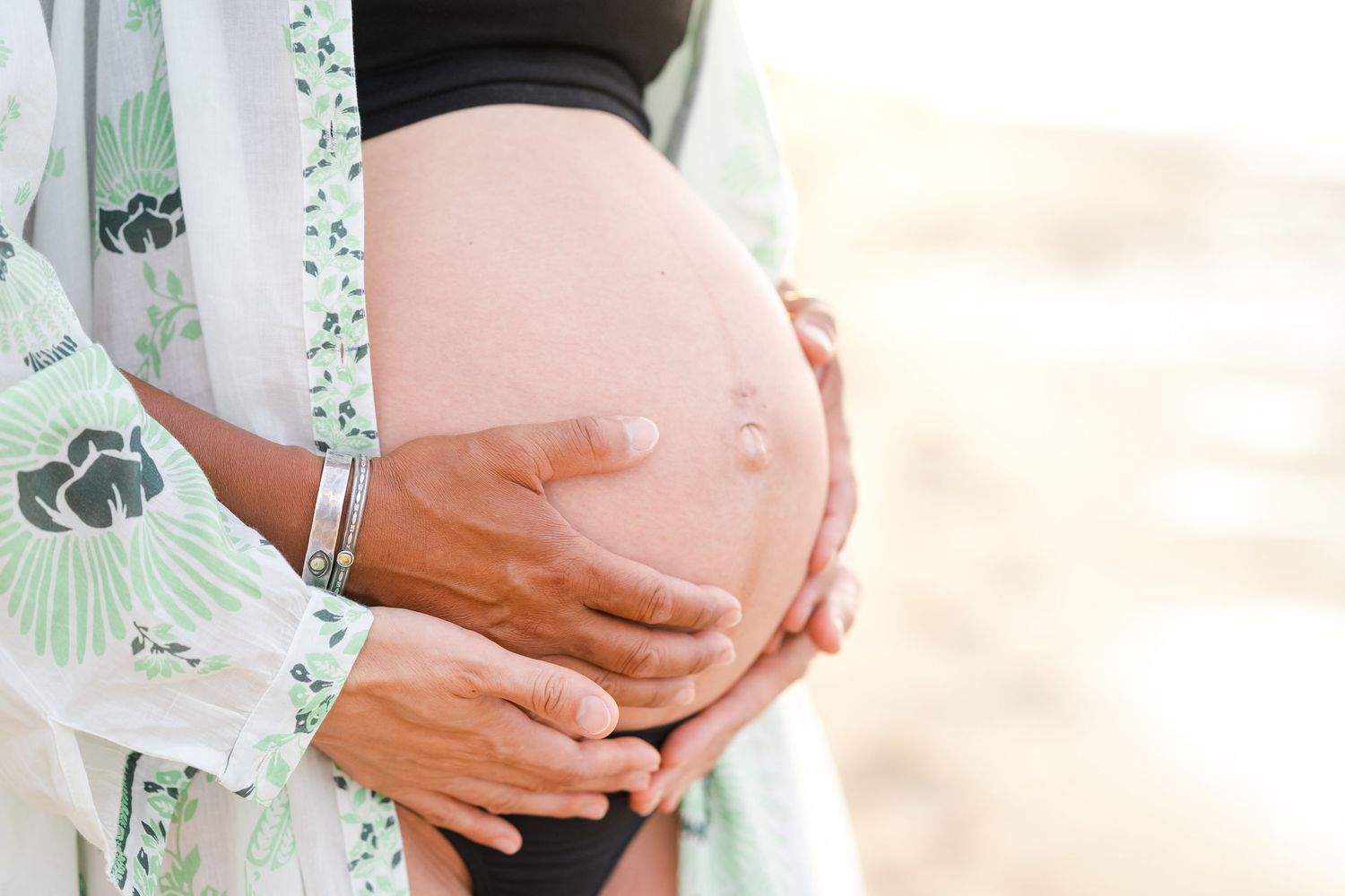 Close-up shot of hands gently cradling a pregnant belly while wearing a mint green floral dress in soft sunlight. Oahu Newborn Photographer