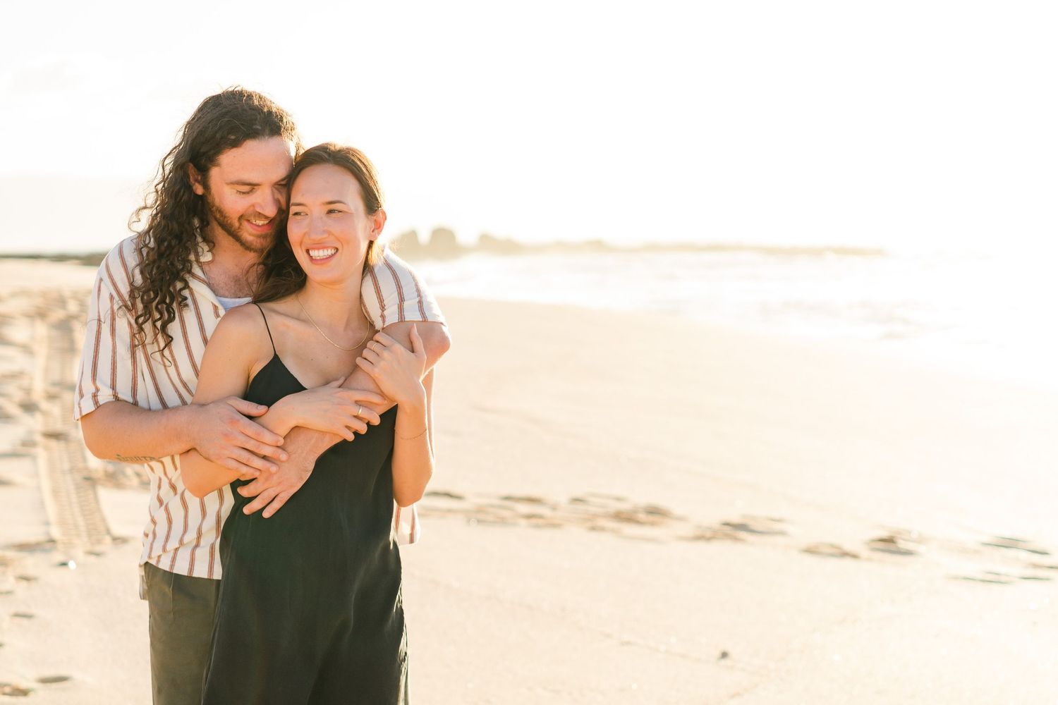 A couple embraces and laughs together on a sunny beach during golden hour.
