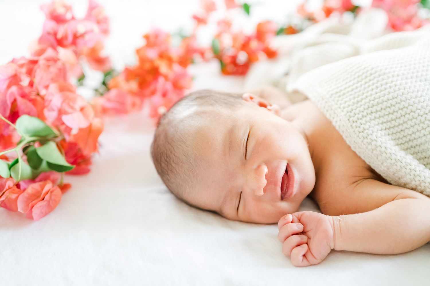 Peaceful sleeping newborn baby wrapped in white blanket lying next to pink coral flowers on white surface. Oahu Newborn Photographer
