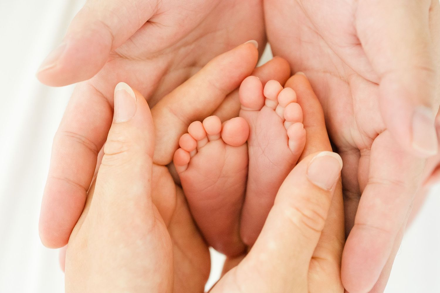 Adult hands gently cradling tiny newborn baby feet against soft pink background. Oahu Newborn Photographer