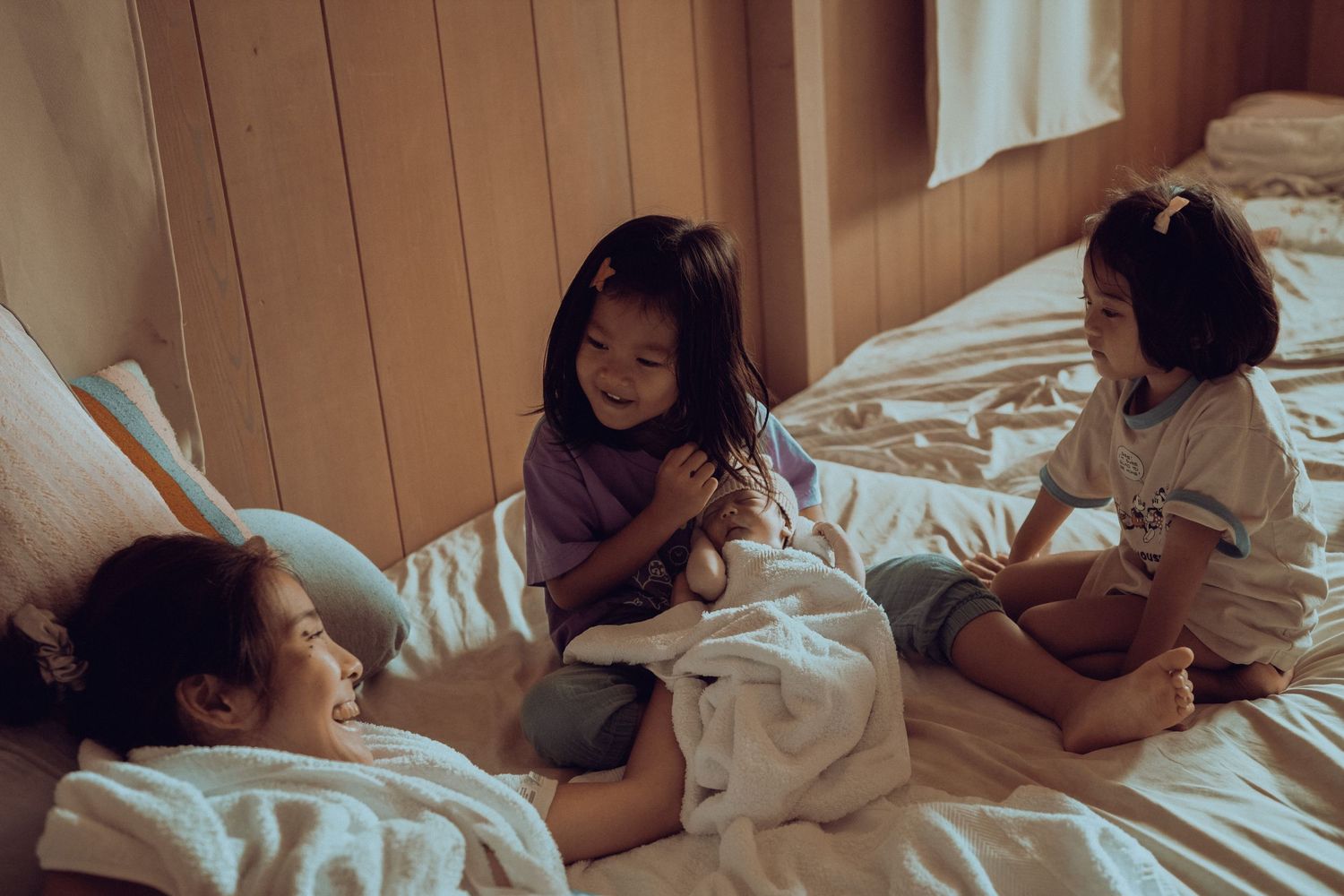 Three children sit together on a bed with white sheets in a cozy room with wood paneled walls.