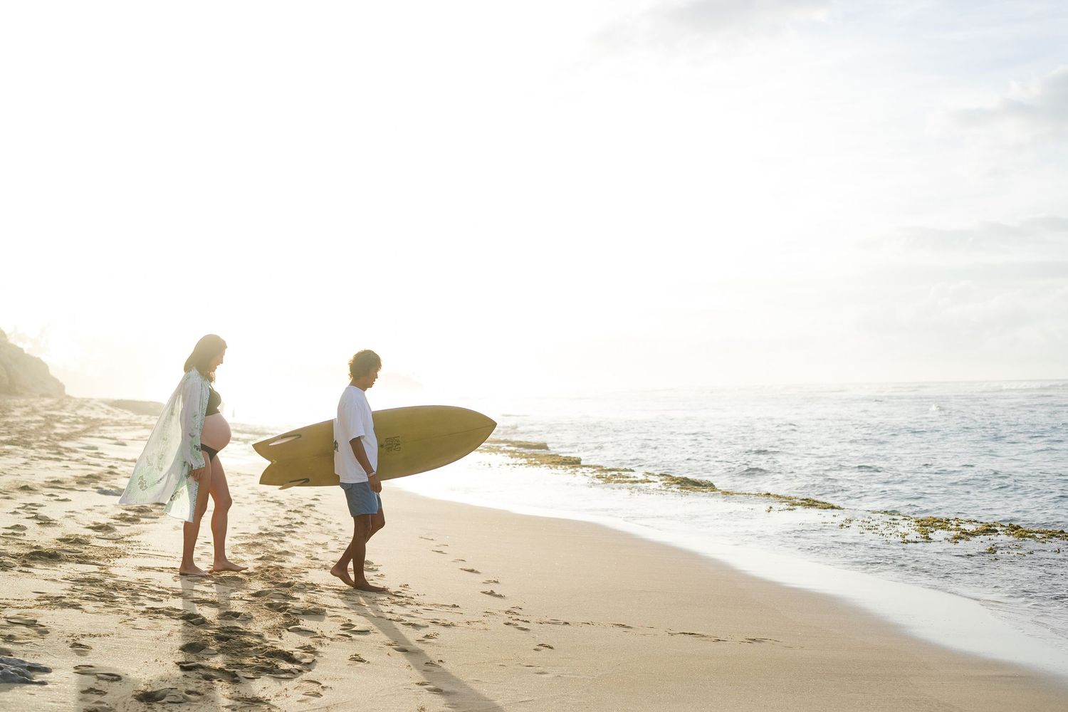 Two surfers walking along a sunny beach with surfboards at sunset while waves crash on the shore. oahu maternity photo