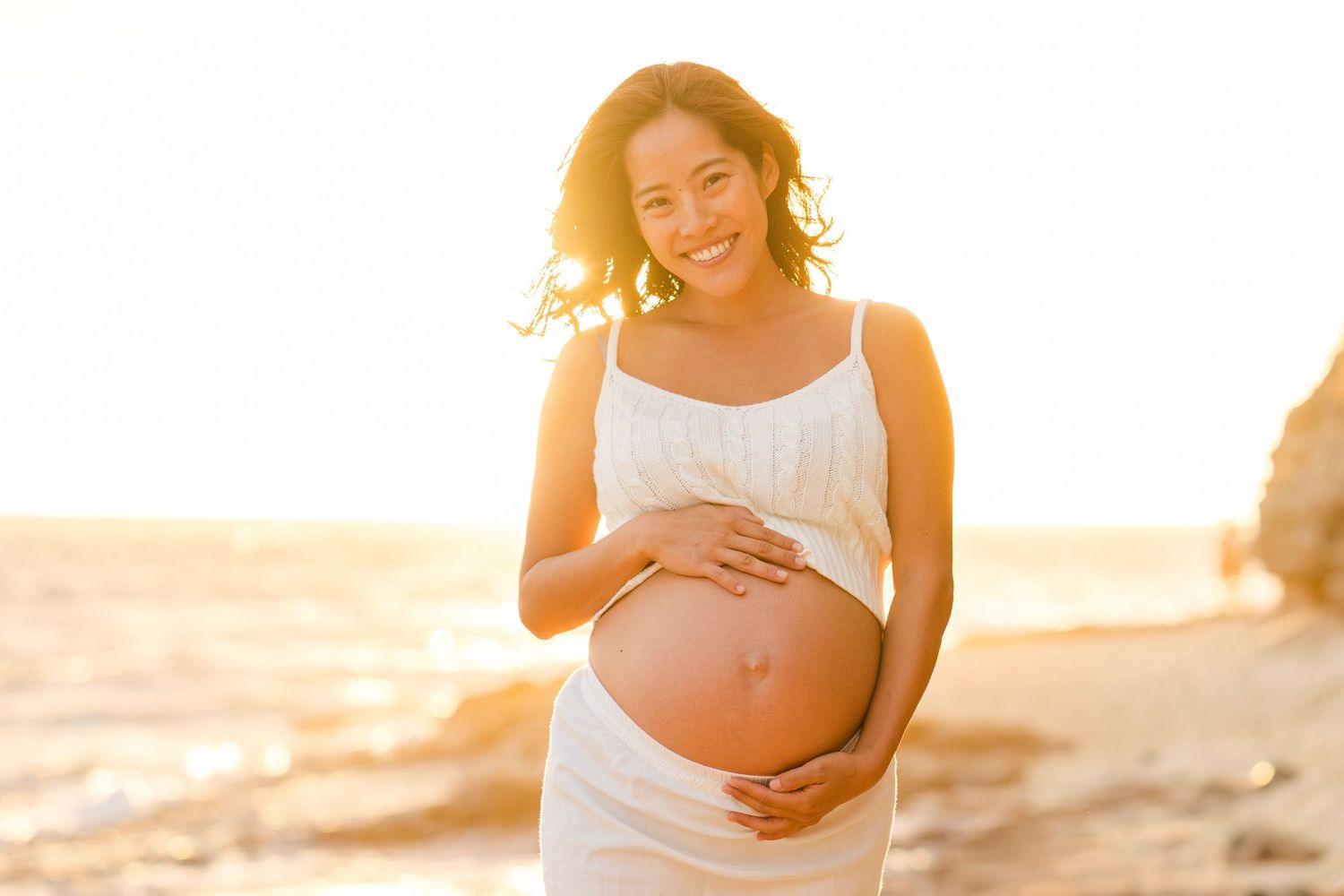 Expecting parent wearing flowing white dress holds bare belly during golden sunset photoshoot on beach.