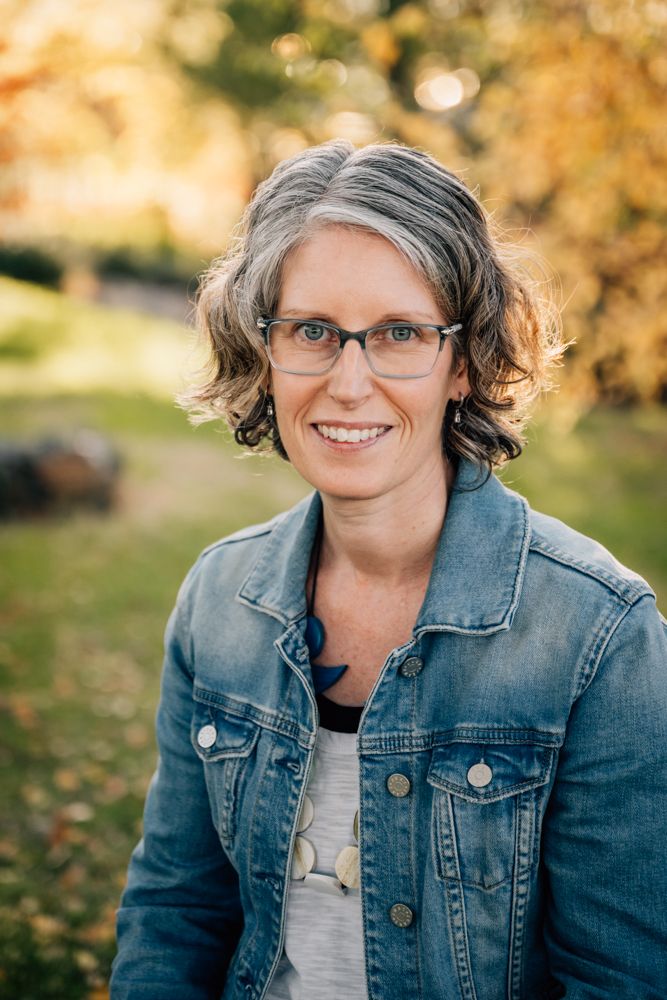 Smiling woman with short gray hair and glasses wearing a denim jacket outdoors in autumn park setting.