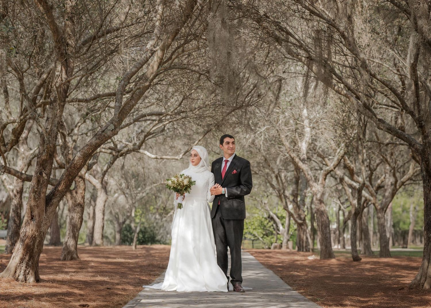 A bride and groom walk together down a path through a tunnel of tall trees in an outdoor forest setting.
