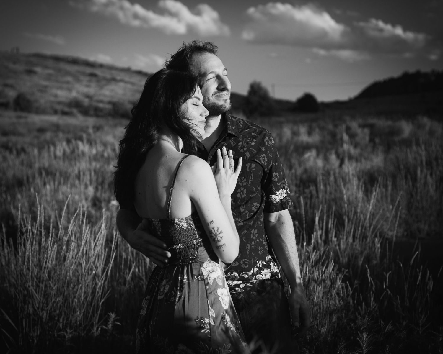 Black-and-white photo of a couple embracing in a field, sunlight creating contrast on the tall grass.