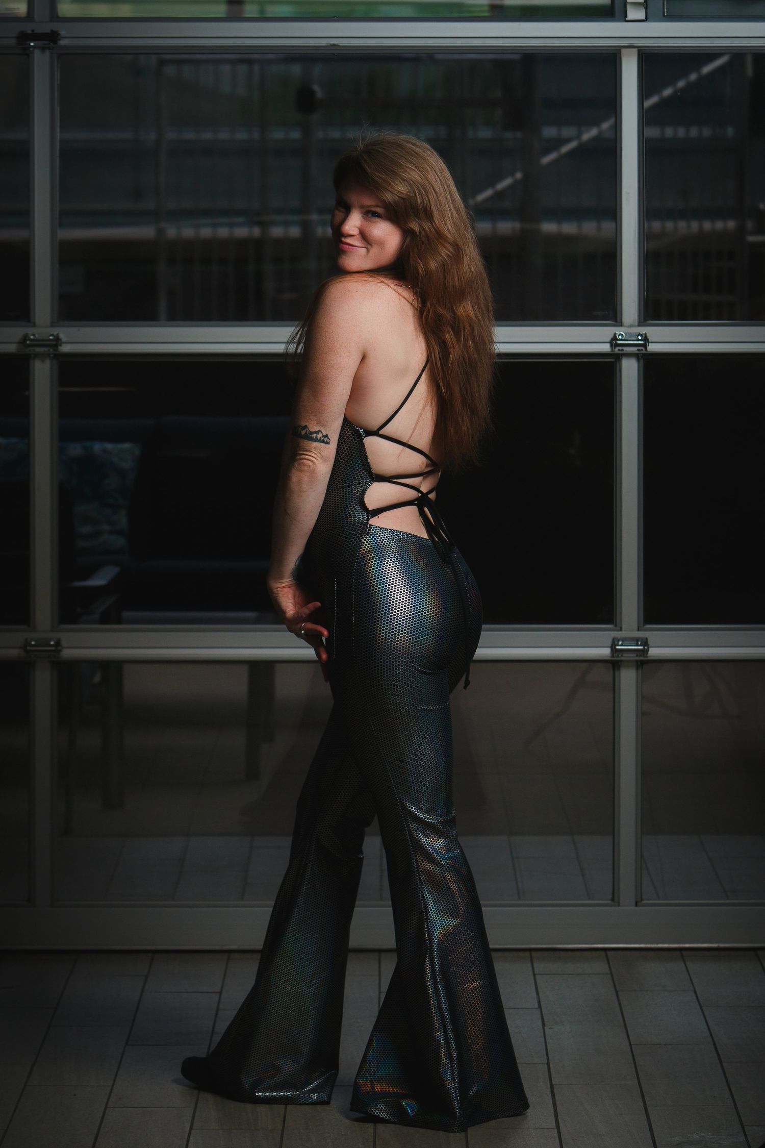 Woman in a shiny black outfit standing indoors in front of glass walls, turned slightly to the side, smiling.