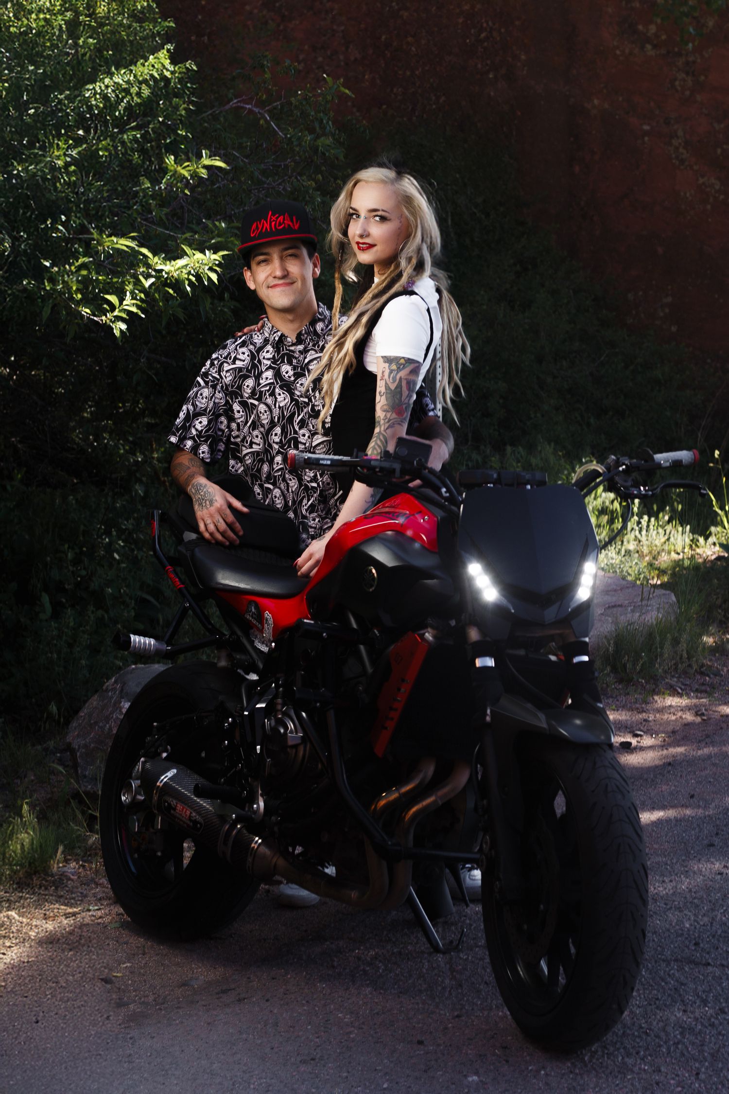 Woman with blonde hair sitting on a motorcycle outdoors, wearing a black vest over white shirt.