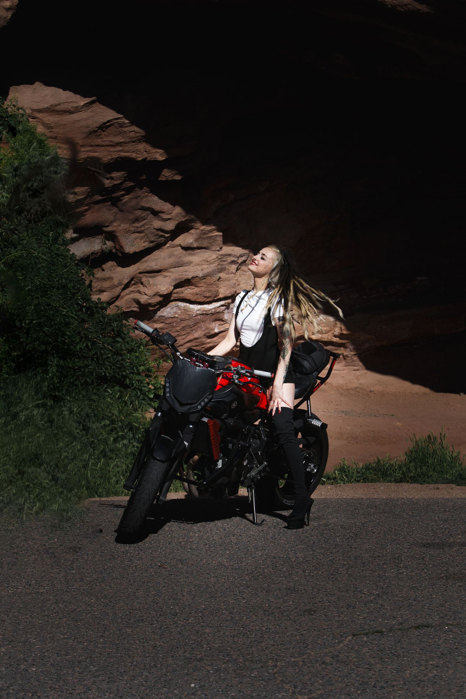 Woman standing next to a black and red motorcycle on a road in front of rocky terrain, looking upward, sunlight highlighting her hair.