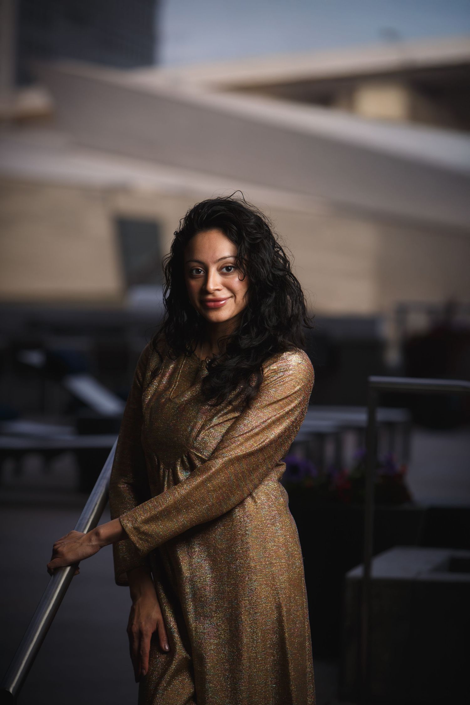 Woman with curly black hair wearing a gold top outdoors in a city setting.