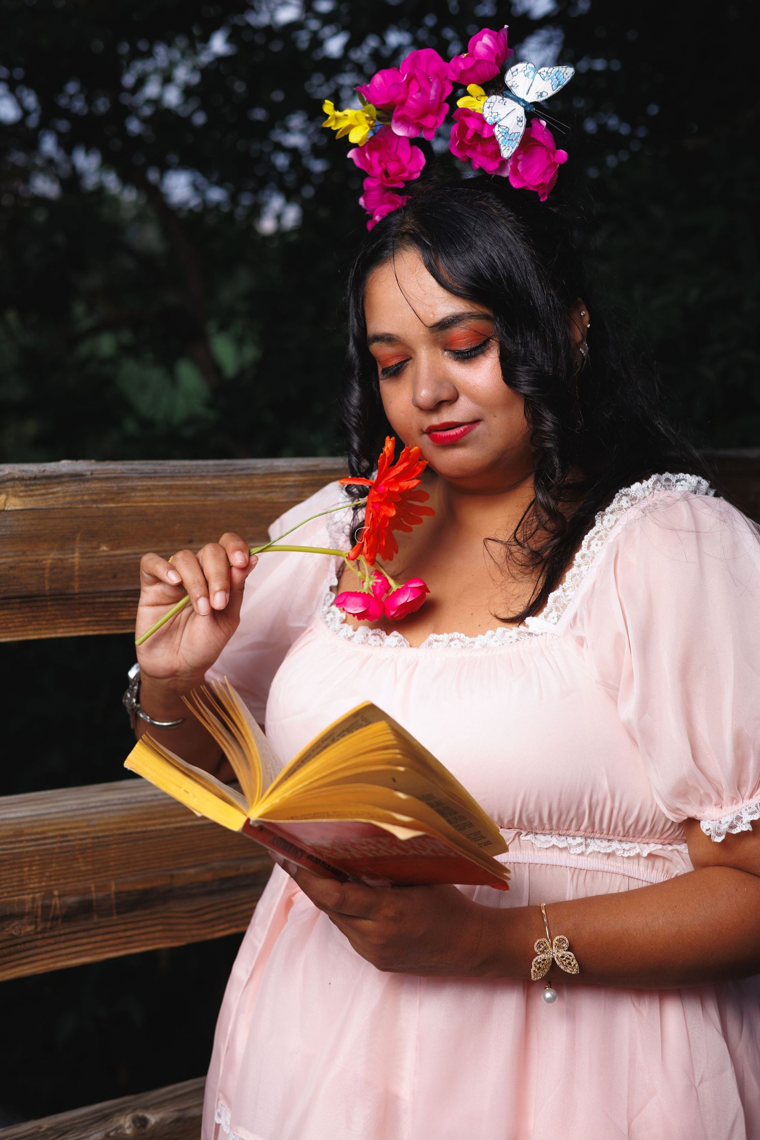 Woman with dark hair wearing a floral headpiece holding a red flower, seated outdoors on a wooden bench.