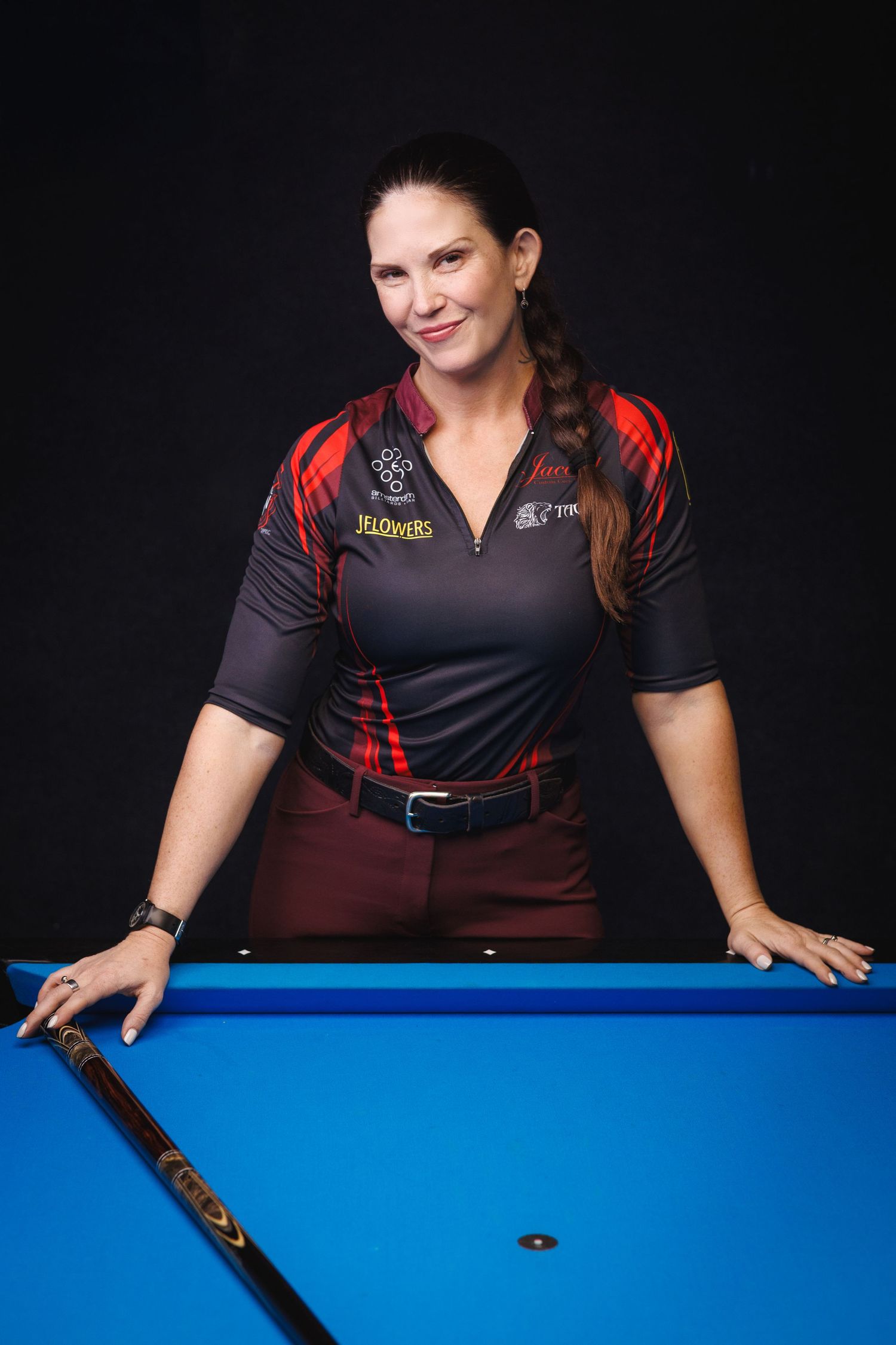 Woman leaning on a blue pool table holding a cue, wearing a black and red sports-style shirt.