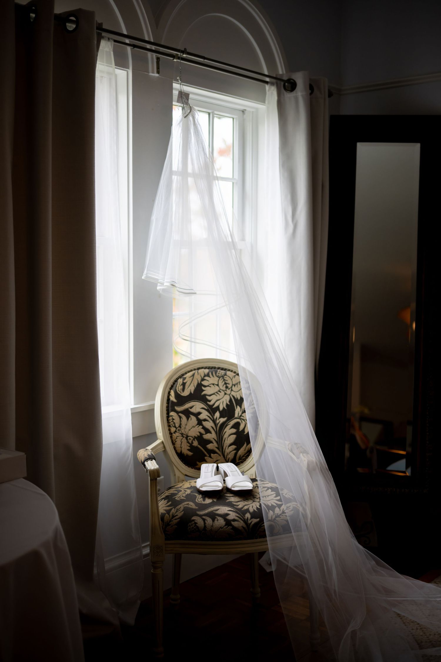 Sheer white curtains billow in the breeze by an antique chair with black floral upholstery near a sunlit window.